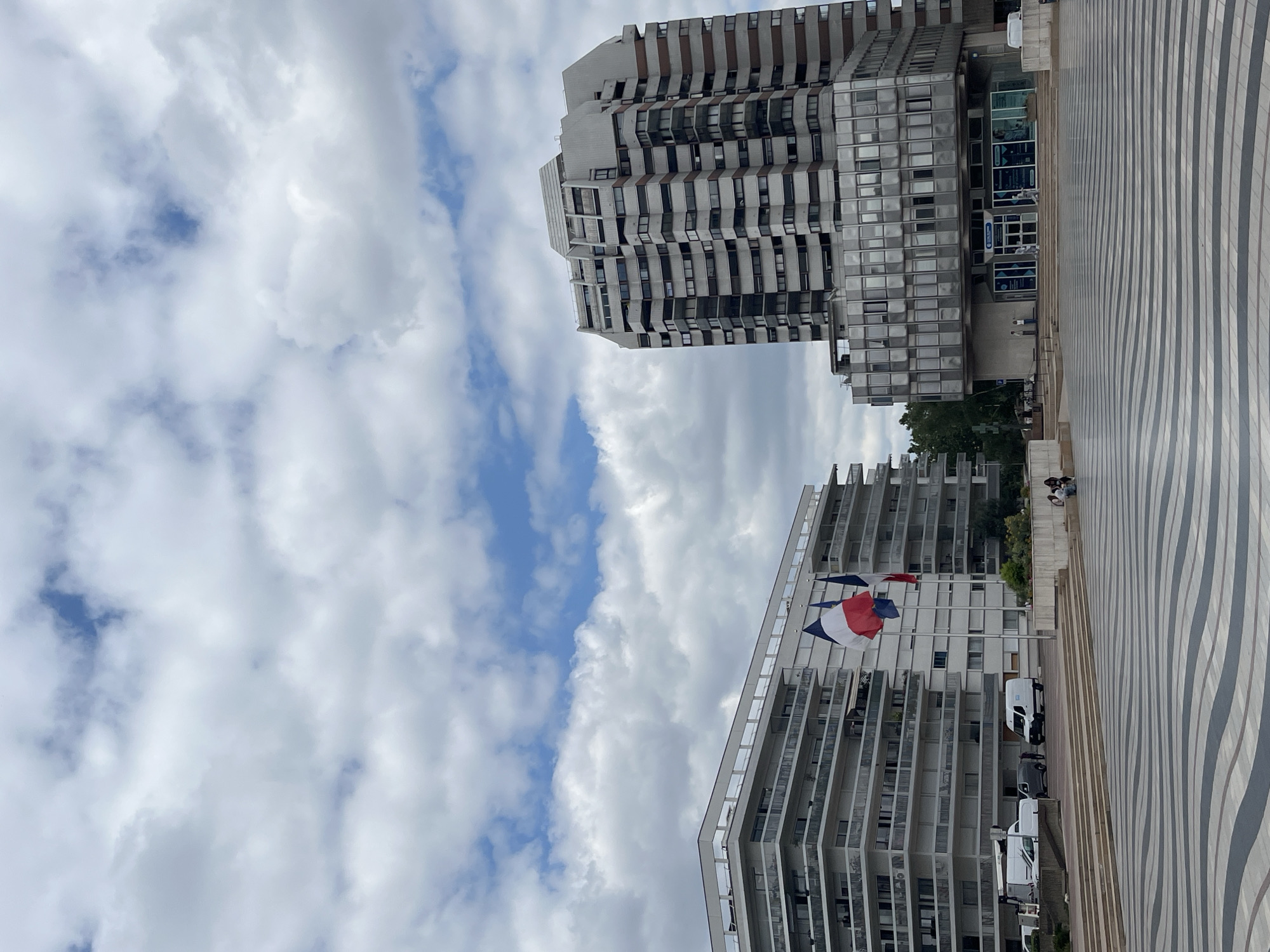 In the city of Créteil, France, a wide, black and white striped plaza occupies the foreground. Beyond the plaza, a modern, multi-story residential building with numerous balconies stands on the left. To the right, a taller, more angular building features a facade of windows and darker horizontal bands. Two French flags are prominently displayed on flagpoles in front of the residential building. Several white vans and one dark colored vehicle are parked near the entrance of the residential building. Further to the right, near the base of the taller building, several people are seated on a raised concrete area. A few individuals are also walking near the entrance of the taller building. The sky above is filled with a mix of white and gray clouds, with patches of blue sky visible.