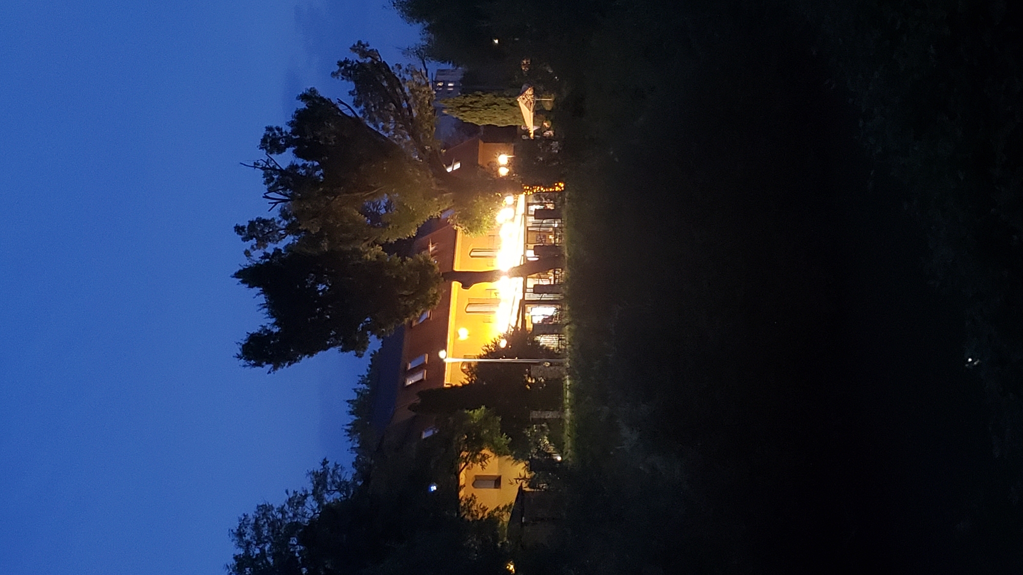 A nighttime, eye-level, vertical shot captures a dimly lit building with lights on inside. The building, a pale yellow structure with a reddish-brown roof, features multiple windows and an illuminated veranda with tables and chairs. The scene is framed by dark trees and foliage in the foreground and background, with the sky a deep, clear blue. The overall ambiance is serene and slightly mysterious. The building is located in Prešov, Slovakia.