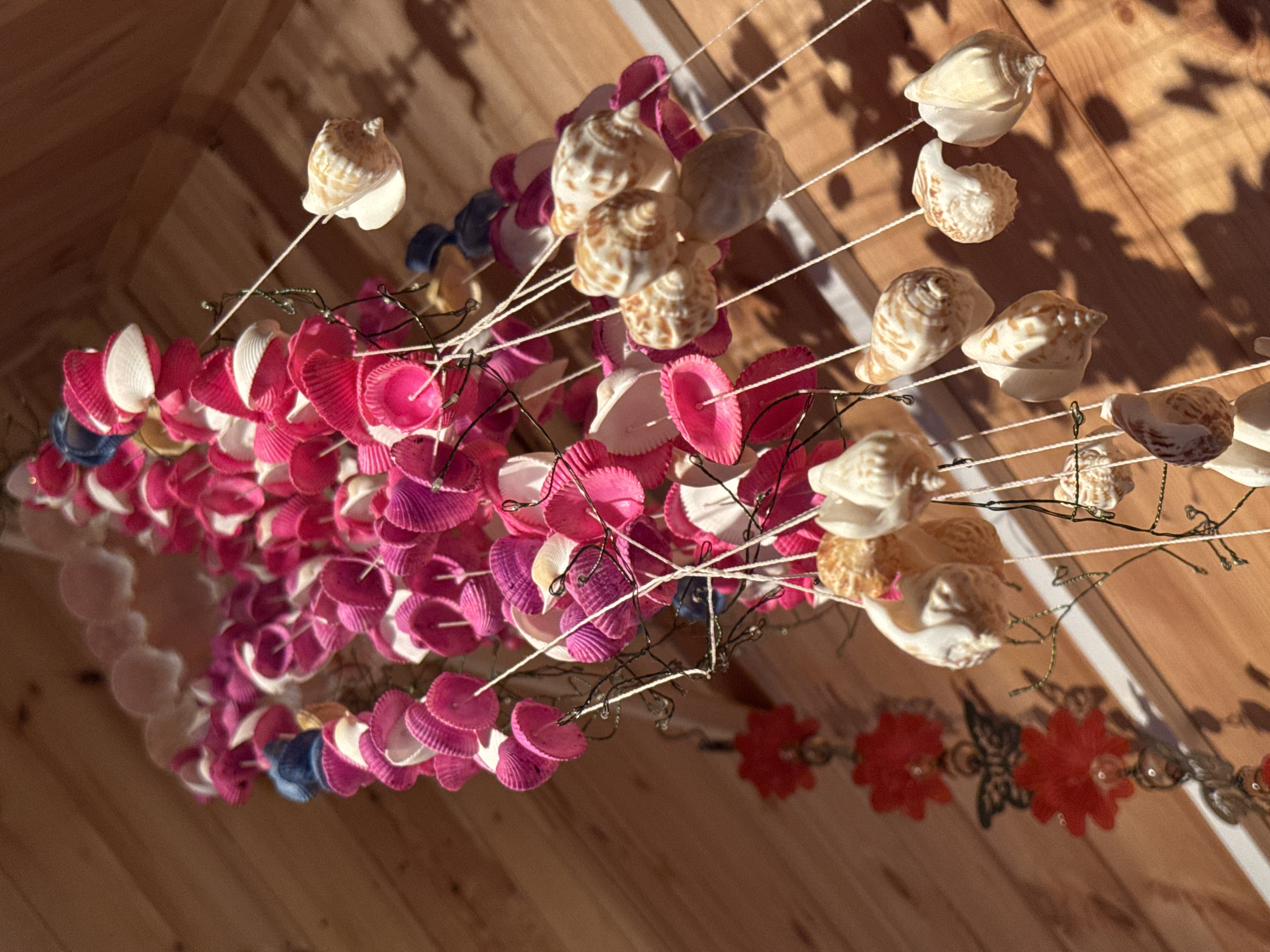 A handmade wind chime crafted from seashells and pink and white shell pieces hangs in front of a wooden wall. The wind chime is made up of multiple strands, each adorned with various shells. Some strands are primarily decorated with pink and white scallop shells, while others feature a collection of natural seashells in various shapes and sizes, with beige and white coloring. There are also decorative elements like red flower shapes and a dark butterfly ornament on one side. The background is a wooden paneled wall, appearing to be an exterior surface due to the sunlight casting shadows. The lighting suggests it is daytime. The location in Zelenaya Roshcha, Russia is not discernible from the image.