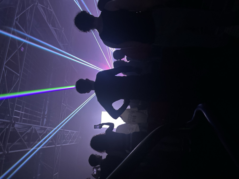 An indoor event scene within the city of Paris, France, depicts a crowd silhouetted against a laser light display. The specific venue within Paris is not visually identifiable. The environment is dark and contains atmospheric haze, through which multiple distinct laser beams in green, purple, blue, and cyan project across the space. Overhead, metallic trusses or a grid ceiling structure are visible. Approximately 10 to 15 human figures are discernible, mostly from the back or in profile, observing the light show. Two prominent figures stand in the immediate foreground, facing away from the viewer; one on the left has hands on hips, and another on the right wears a sleeveless top. To the left, a person with curly hair holds a smartphone vertically, and another individual further left holds a phone horizontally, both actively recording the event. A dark, curved railing is present in the lower-left foreground.