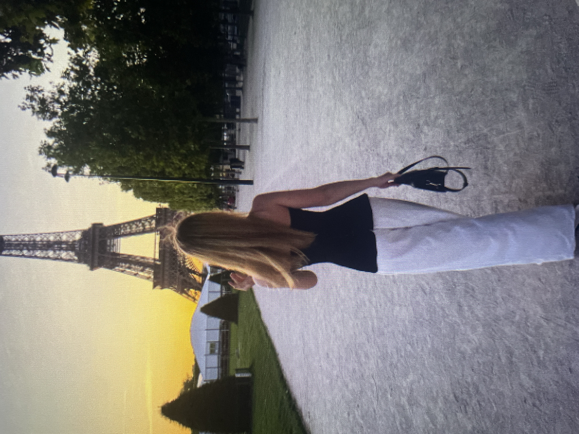 The image depicts a woman from behind walking away from the camera towards the Eiffel Tower in Paris, France. She has long, blonde hair and is wearing a black strapless top and white wide-leg pants. She is holding a small black purse in her right hand. The Eiffel Tower is visible in the background, silhouetted against a yellow and orange sunset. The scene appears to be a park or promenade, with trees lining the path and the iconic landmark prominently featured. There are no elements in the image that suggest the location is in Kołobrzeg, Poland.