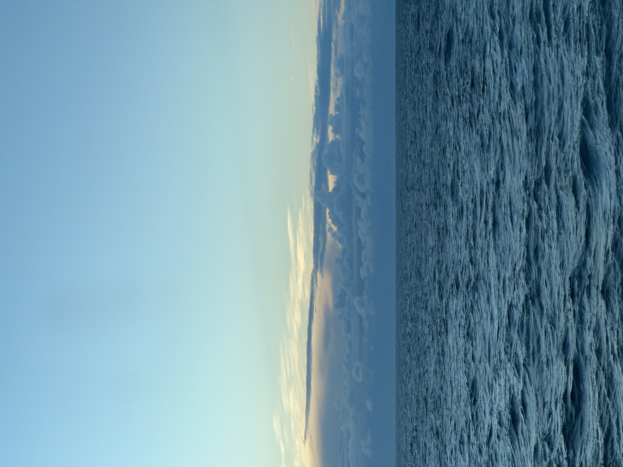 The image depicts a calm ocean scene under a pale blue sky. The water is a deep blue with small waves and ripples, indicating a gentle surface. A band of clouds, tinged with yellow and pink from what appears to be the setting or rising sun, stretches across the horizon. The sky above the clouds is a clear, gradient blue, fading from a lighter hue near the horizon to a deeper shade at the top of the frame. The scene is entirely natural, with no man-made structures or living beings visible. The location is not specified within Poland, or any city.