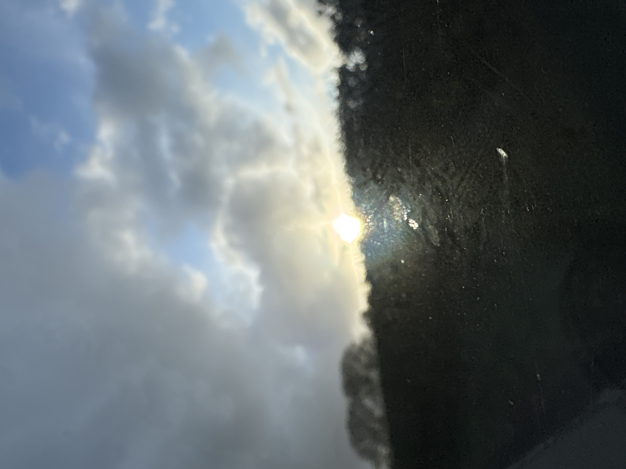 The image shows a view of clouds and sunlight partially obscured by a dark, possibly dirty, surface. It is unclear whether the dark surface is a window or another object. The sky is partly cloudy with patches of blue visible. The sun shines through a break in the clouds. The location is impossible to determine from the image provided. No people, subjects, objects, actions, or interactions are visible. The environment consists of the sky and an unclear dark surface.