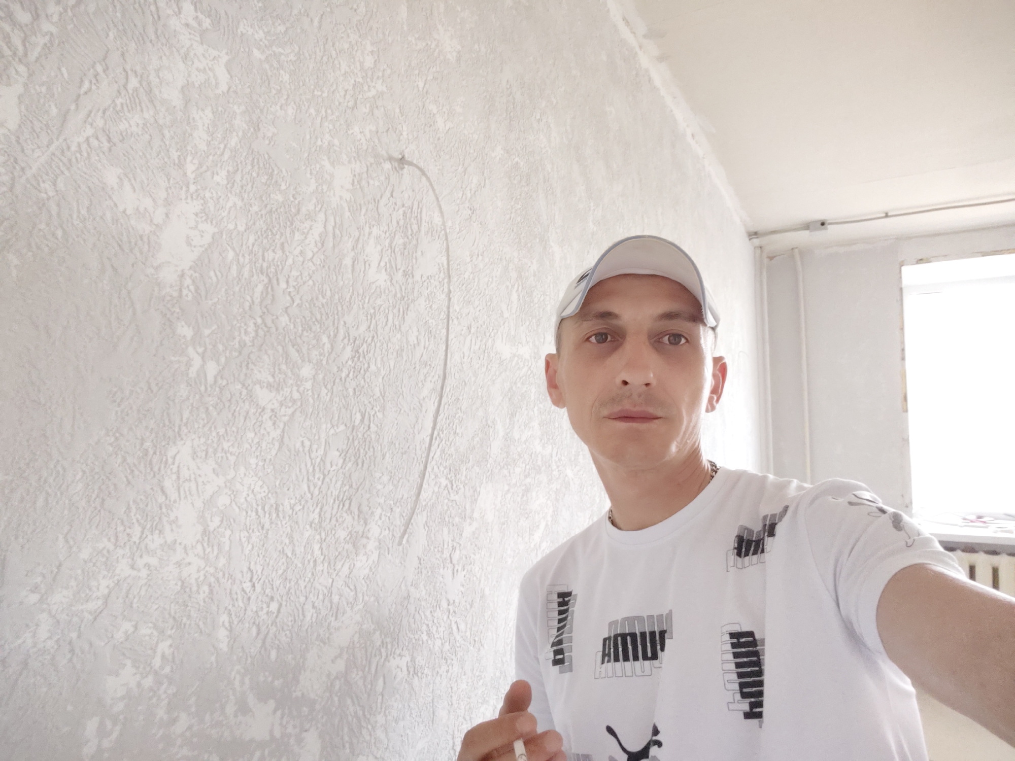 The image shows a man wearing a white Puma t-shirt and a white baseball cap, taking a selfie in what appears to be a room undergoing renovation or construction. The walls have a textured, off-white finish, and there is a faint, curved line drawn on the wall to the left of the man, possibly a marking for work. The ceiling is also visible, with some electrical conduit. To the right, a window with bright light coming through is partially visible, along with a radiator. The man is holding a cigarette in his right hand. The scene is indoors, likely residential, and the presence of construction materials or unfinished work suggests it could be in the city of Bor, Russia, as indicated by the context.