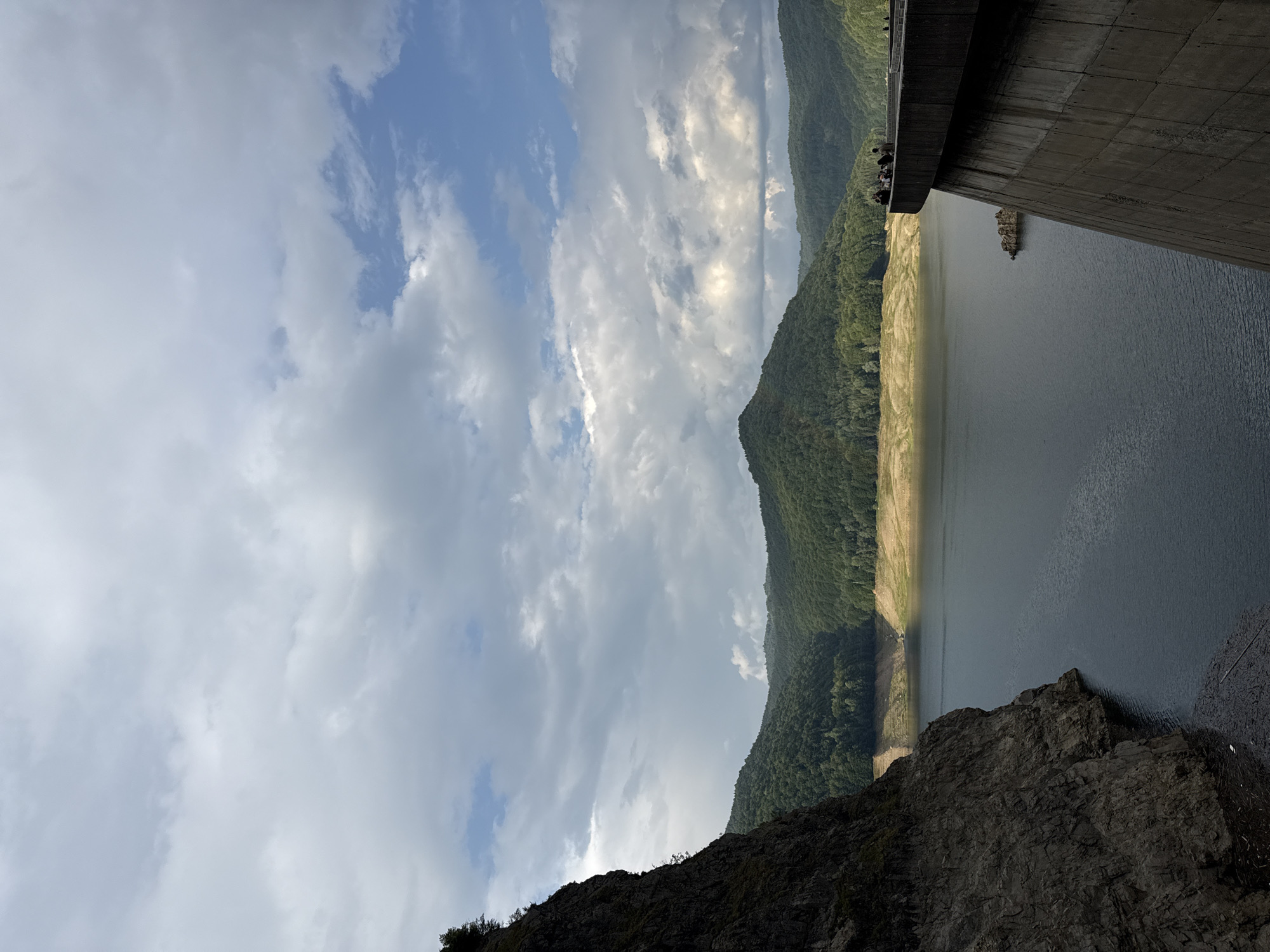 The image depicts the Vidraru Dam, a concrete arch dam situated on the Argeș River in Romania, near the city of Arefu. The dam is a prominent structure, with its massive concrete face dominating the foreground on the right side of the frame. On top of the dam, a group of approximately ten individuals are visible standing and observing the landscape, with one person appearing to lean over the edge. The reservoir behind the dam is a large body of dark water, reflecting the cloudy sky. The surrounding environment consists of lush, green, forested hills that rise steeply from the water's edge. A large, dark, rocky cliff face is present on the left side of the image, creating a dramatic contrast with the smooth concrete of the dam and the softer forms of the hills. The sky above is filled with a mix of white and grey clouds, with patches of blue sky visible, suggesting a dynamic weather pattern. Debris, such as branches and leaves, are visible floating on the water near the base of the cliff. The scene conveys a sense of scale and the imposing nature of the dam against the natural landscape.
