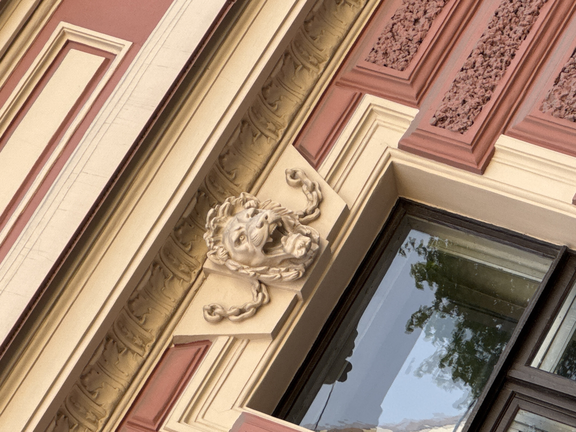An angled, close-up shot captures a section of an ornate architectural facade, likely from a building in Saint Petersburg, Russia. The building features a multi-layered design with prominent cream or beige elements framed by reddish-brown sections.
A central feature is a sculpted lion's head, made of a light-colored material, possibly stone or stucco, positioned on a cream-colored block. The lion is depicted roaring, with an open mouth, prominent fangs, and expressive eyes. Its mane or beard appears to be stylized as flowing chains, adding to its imposing look.
Above and to the side of the lion sculpture, decorative elements include a patterned frieze with a swirling, organic motif in a similar cream/beige tone, and rectangular panels filled with a rough, granular texture in the reddish-brown color. The intricate layering of cornices and moldings creates depth and visual interest.
Below the lion sculpture and extending to the right, a dark-framed window reflects the outside world. The reflection clearly shows the canopy of trees and a bright sky, suggesting a daytime scene. The window frame is dark, contrasting with the lighter tones of the surrounding facade. The overall impression is one of classical or historical European architecture, characteristic of a city like Saint Petersburg. No discernible text is visible in the image.
