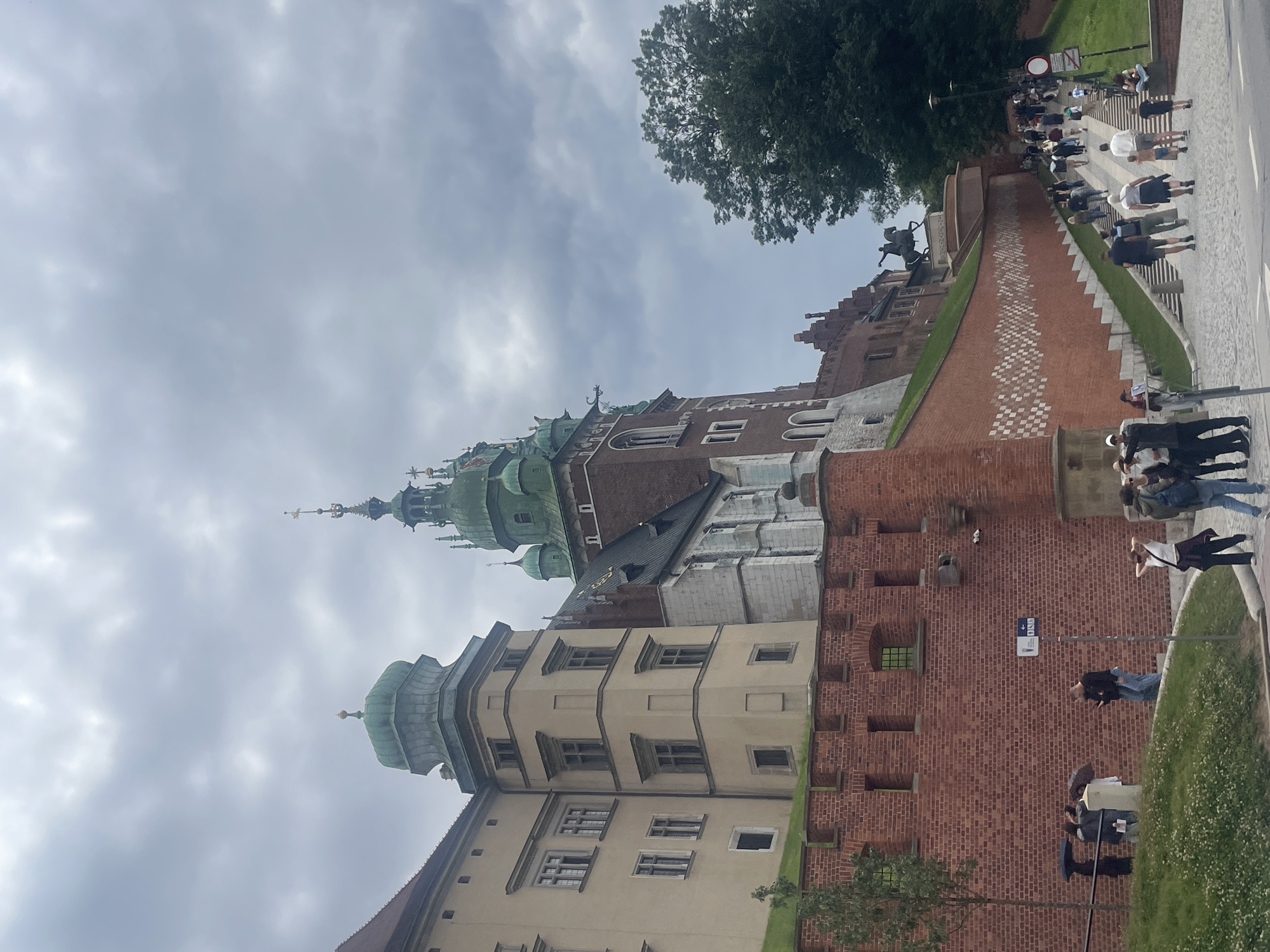 The image depicts the Wawel Castle in Krakow, Poland, on an overcast day. The castle features a prominent brick wall with crenellations and several windows. To the left, a pale yellow building with multiple windows rises. Adjacent to it, a larger structure with a green dome and a tall, green-roofed tower dominates the skyline. In the foreground, a stone pathway leads up a wide staircase lined with a brick wall on the left and a grassy slope on the right. Numerous people are walking up the stairs, some individually and some in small groups. A bronze equestrian statue is visible at the top of the stairs, positioned against the backdrop of lush green trees. To the left of the stairs, near the brick wall, a few individuals are standing under umbrellas.