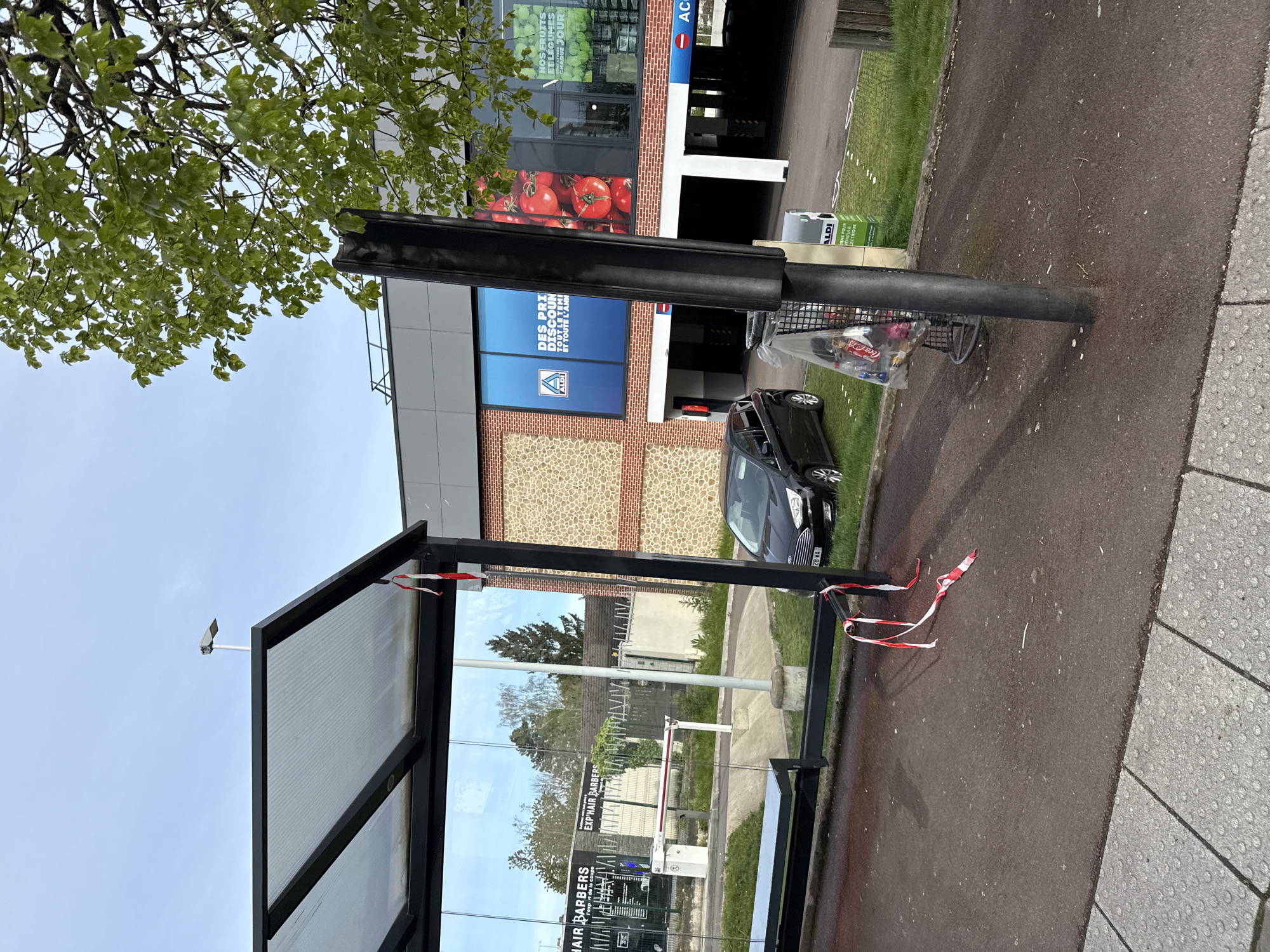 The image shows an Aldi store in Domont, France. A bus shelter stands on the pavement outside the store, partially obstructed by red and white striped tape. A black car is parked next to the bus shelter. In the background, a tree partially obscures the view of the building. A shopping basket filled with garbage is attached to a pole.
