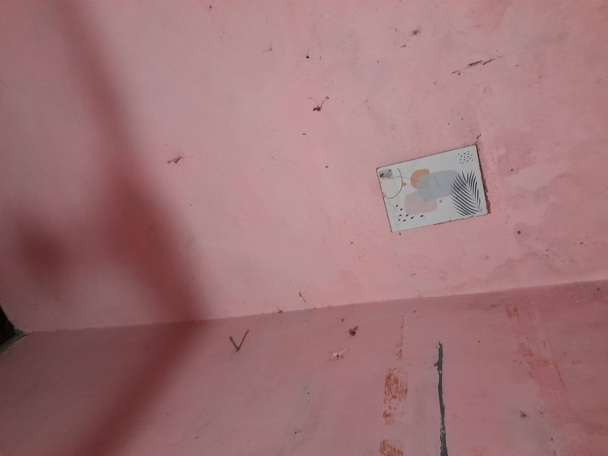 A pink wall in a room. The wall has some small dark spots and a few scratches. In the upper right quadrant of the image, a small decorative tile is attached to the wall. The tile has an abstract design with cream, gray, and orange shapes, along with some black dots and a drawing of a palm leaf. In the lower left quadrant, there is a subtle shadow cast across the wall, suggesting a light source from the left. A faint "V" shape is also visible on the wall in the lower middle section. The overall impression is of a simple, domestic interior. There is no discernible time of day, weather, or emotional cues. There is no visible text.