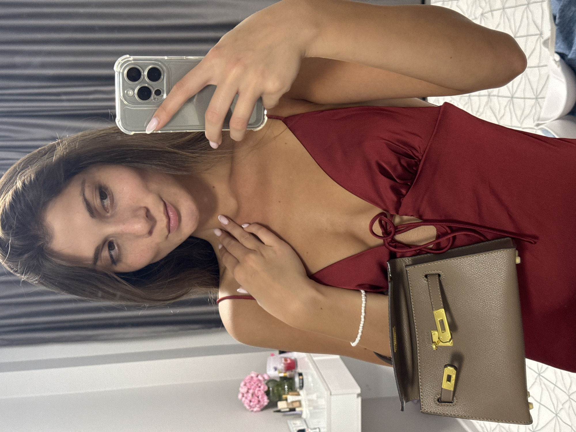 A young woman is taking a selfie in what appears to be a bedroom. She is wearing a dark red satin slip dress and holding a small taupe handbag with gold hardware. She is also wearing a delicate pearl bracelet on her left wrist. The background shows a vanity with some makeup products and a vase of pink flowers, and a dresser with a mirror. The lighting suggests it is daytime. The image does not provide any information to determine the location in Burgas, Bulgaria.