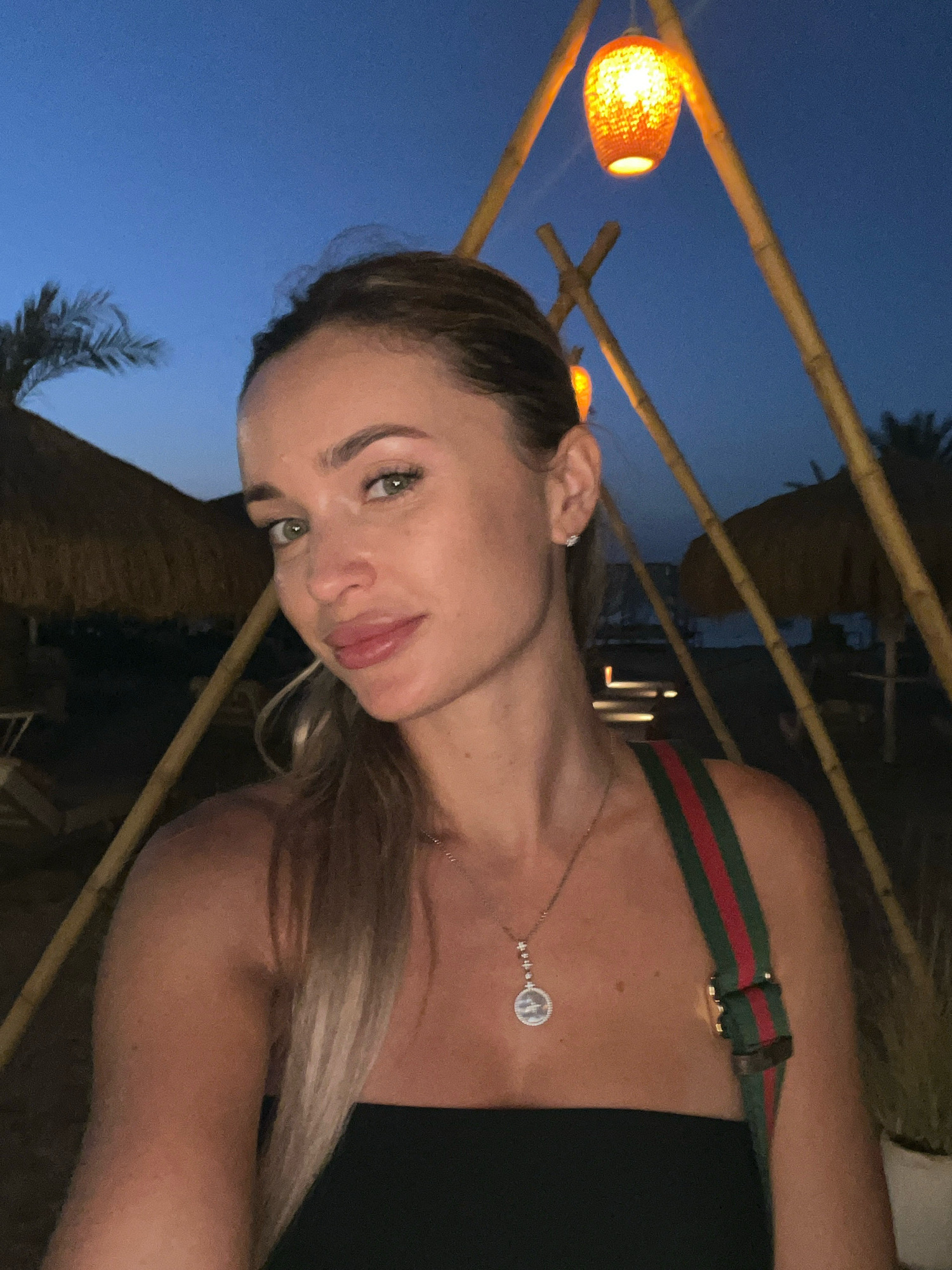 A woman is captured in a selfie at dusk in Sharm El Sheikh, Egypt. She has light skin, green eyes, and blonde hair pulled back. She is wearing a black strapless top and a necklace with a circular pendant. A green and red striped strap of a bag is visible over her shoulder. Behind her, bamboo poles form a triangular structure, with an orange lantern hanging from it. The background shows a thatched-roof structure and palm trees against a deep blue sky. The scene suggests a relaxed, outdoor setting, possibly a resort or beachside cafe.