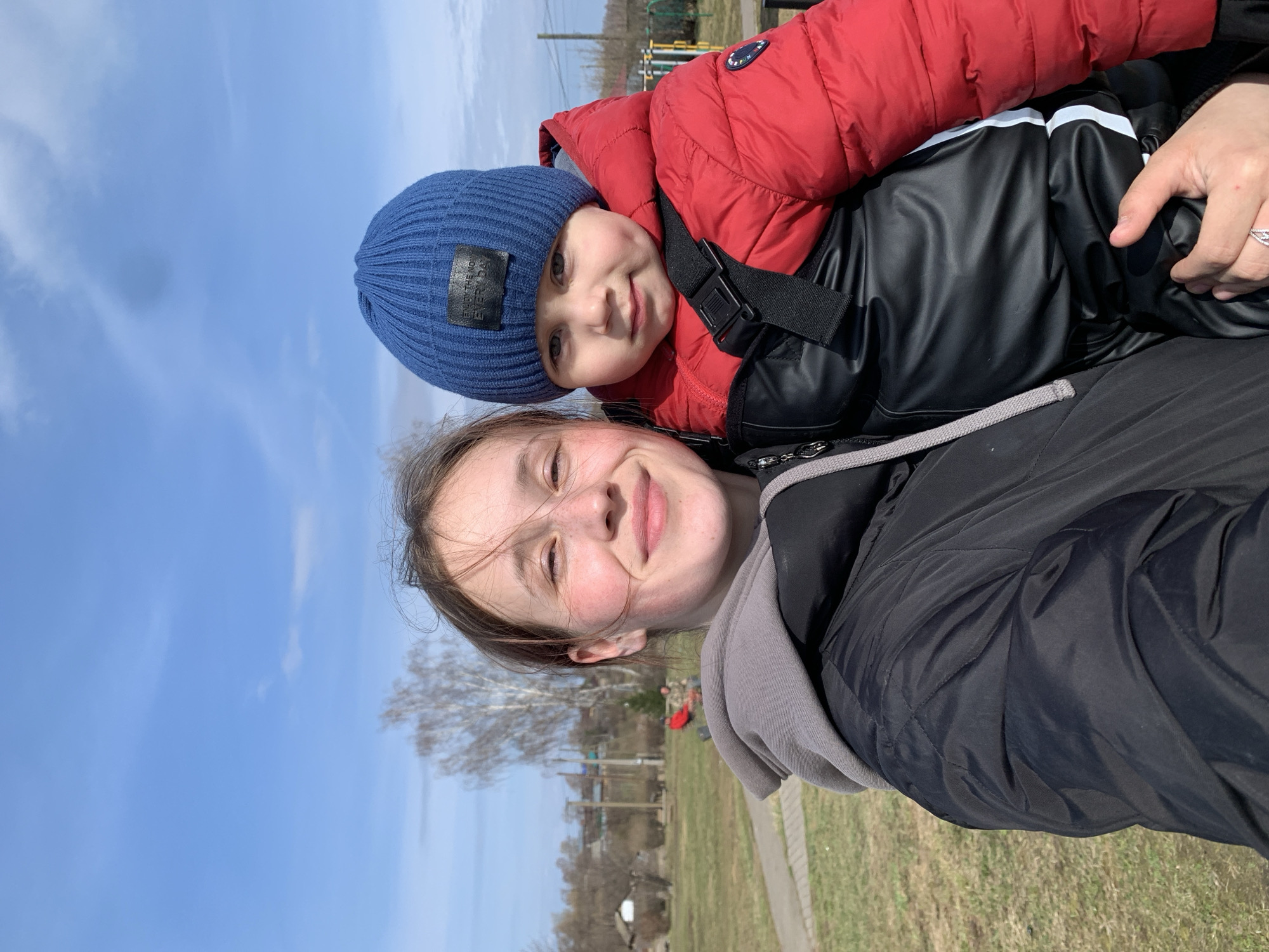 A woman is holding a young child outdoors. The child wears a blue knit cap and a red puffer jacket. The woman wears a black jacket with a gray hood. In the background, there's a grassy area, bare trees, a structure resembling playground equipment, and a blue sky. The photo was taken in Фоки, Russia.