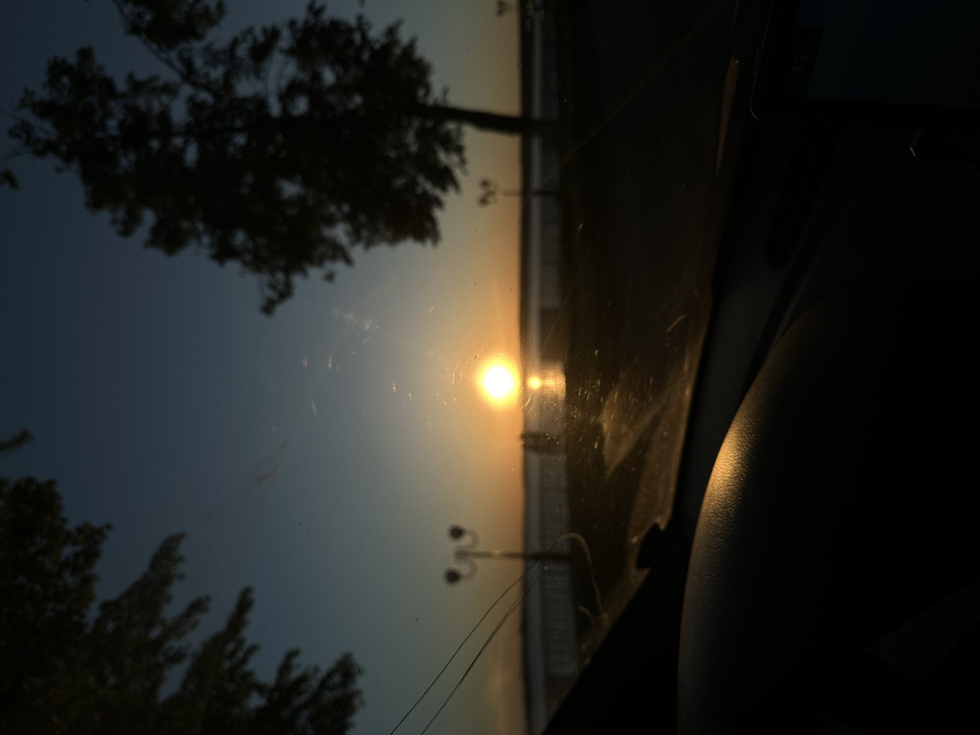 The image appears to have been taken from inside a vehicle, looking out towards a sunset or sunrise. The sun is positioned in the center of the frame, obscured by the horizon, which suggests it is setting or rising. There are silhouettes of trees and streetlights on either side of the horizon.
It is impossible to definitively ascertain the exact location in Votkinsk, Russia, as no specific identifiable landmarks are visible. The image shows a coastline with an area of city infrastructure, which suggests it might have been captured near the Votkinsk Reservoir (part of the Kama River) or the Votka River.
The interior of the vehicle is dimly lit, obscuring many details. Part of the dashboard and what appears to be a mobile device or navigation system are visible. The windshield of the vehicle is dirty, with smudges and streaks visible.