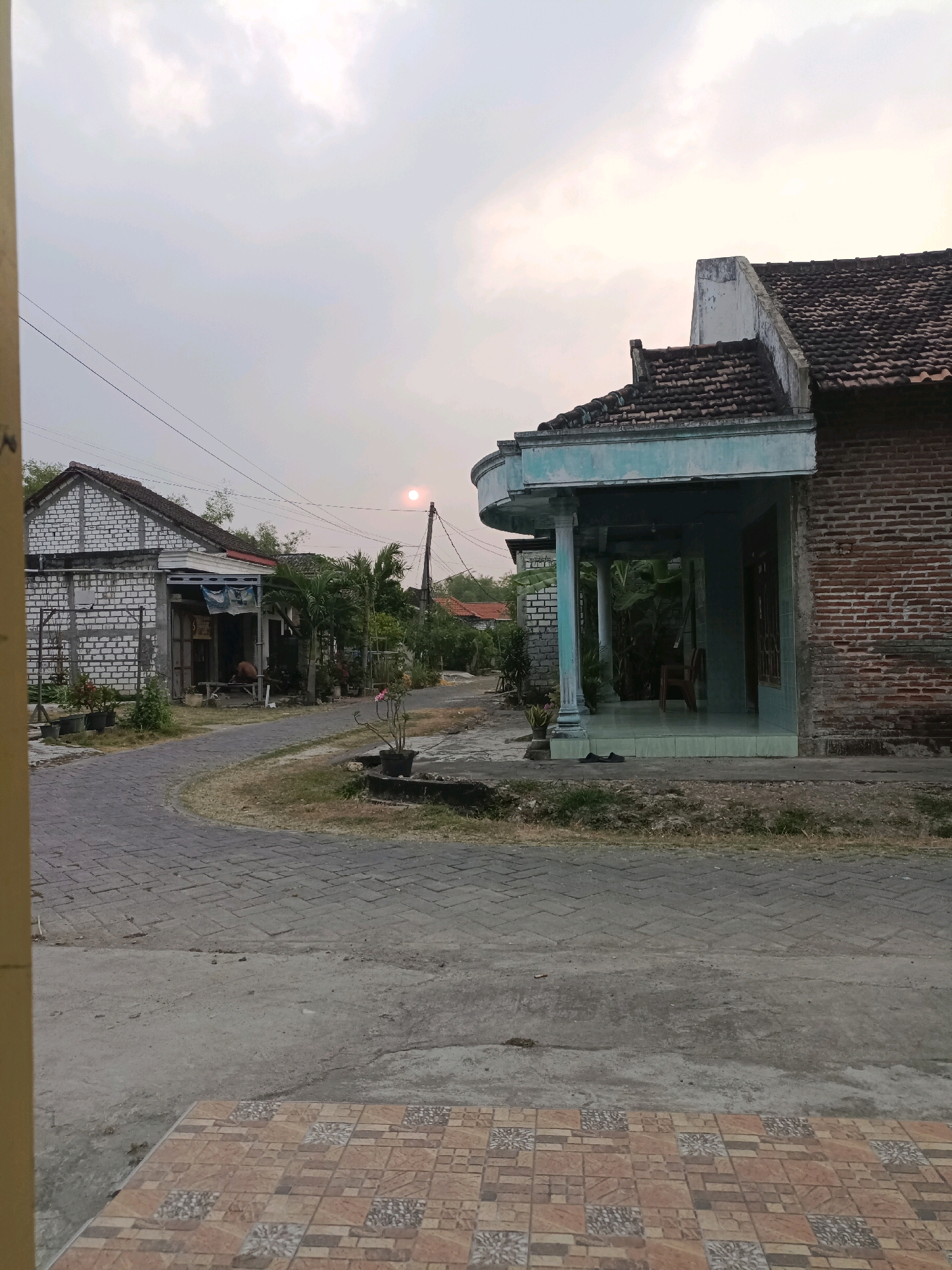 The image depicts a street scene in Rancang Kencono, Indonesia, under a hazy sky with the sun low on the horizon, possibly during sunrise or sunset. The perspective is from ground level, looking down a paved road that curves to the left. On the right side of the road is a brick house with a tiled roof, featuring a covered porch with light blue trim and columns. A pair of dark sandals are placed on the porch. Adjacent to this house, the road continues past another structure built with white bricks, which has an open front area with some shelving and a person, appearing to be a male, sitting on a wooden structure. This person is partially visible and appears to be working or resting.
Further down the road, a small pink flower in a pot is situated on the left side of the pavement, near the brick house. The road is paved with interlocking concrete blocks in some sections and asphalt in others. Vegetation, including palm trees and other plants, is visible throughout the scene, lining the road and around the houses. Electrical wires are strung across the sky, supported by utility poles. The overall impression is of a quiet, residential street in a tropical or subtropical climate, with the natural light suggesting it is either early morning or late evening.