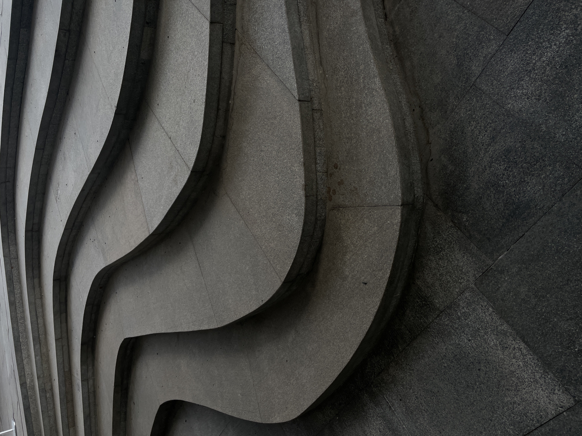 This is a close-up, slightly angled shot of an architectural feature, specifically a multi-tiered, wave-like structure made of grey stone tiles. The tiers curve and undulate, creating a sense of depth and rhythm. The tiles have a mottled, granular texture, with subtle variations in tone across their surfaces. Dark shadows accentuate the recesses between the tiers, enhancing the contrast and highlighting the form. There are seams apparent in the tiles. The location is not specified, and therefore there is insufficient information to determine the location as Moscow, Russia. There are no people visible in the image.