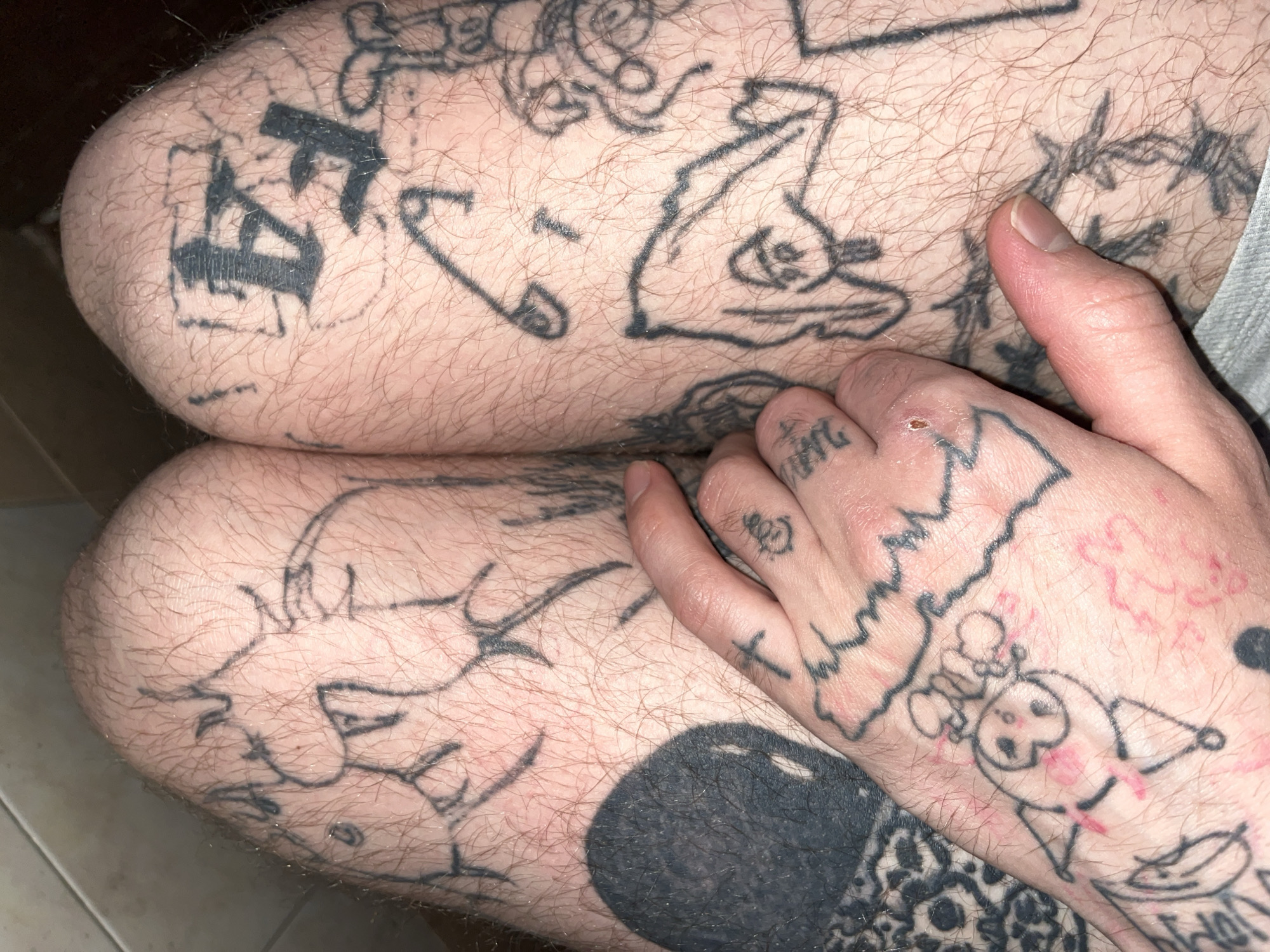 The image is a close-up, high-angle shot of a person's legs and hand, adorned with numerous black ink tattoos. The skin appears to have light hair. The tattoos are varied in style and subject matter, including what appear to be cartoonish figures, letters, and abstract designs. One tattoo on the hand depicts a crescent moon shape with facial features and an object resembling a halo or crown. Another tattoo on the leg shows a large, dark circular shape. A hand is visible in the foreground, resting on the leg, also tattooed with small designs, including a cross. The environment appears to be indoors, possibly a bathroom, indicated by the tiled wall in the upper left corner. The lighting is moderate, casting some shadows. No city or specific location information is discernible from the image.