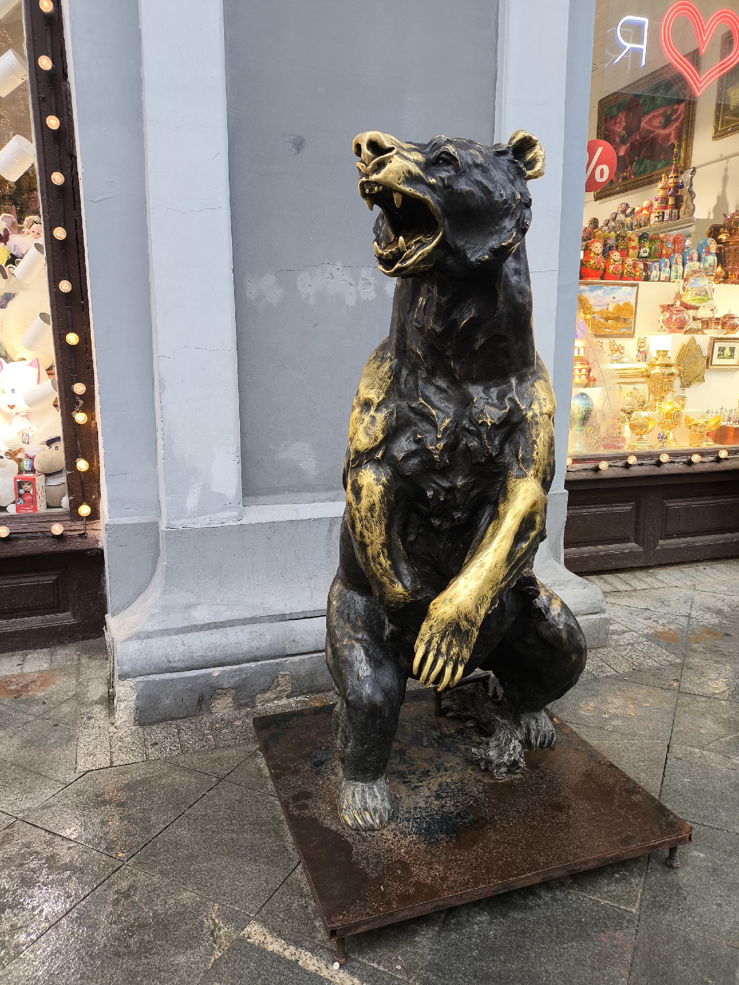 A bronze and gold bear statue sits on a rusty brown metal platform in front of two store windows on a wet, tiled city street. The bear is in a seated posture, appearing to roar with its mouth open. The exterior walls of the building are a light grey. The left store window displays stuffed animals and is framed with lights. The store window on the right contains various decorative items, including Matryoshka dolls, set against a dark wooden frame. A neon sign with a heart is visible in the reflection of the window. The location is identified as the city of Unknown, Unknown.