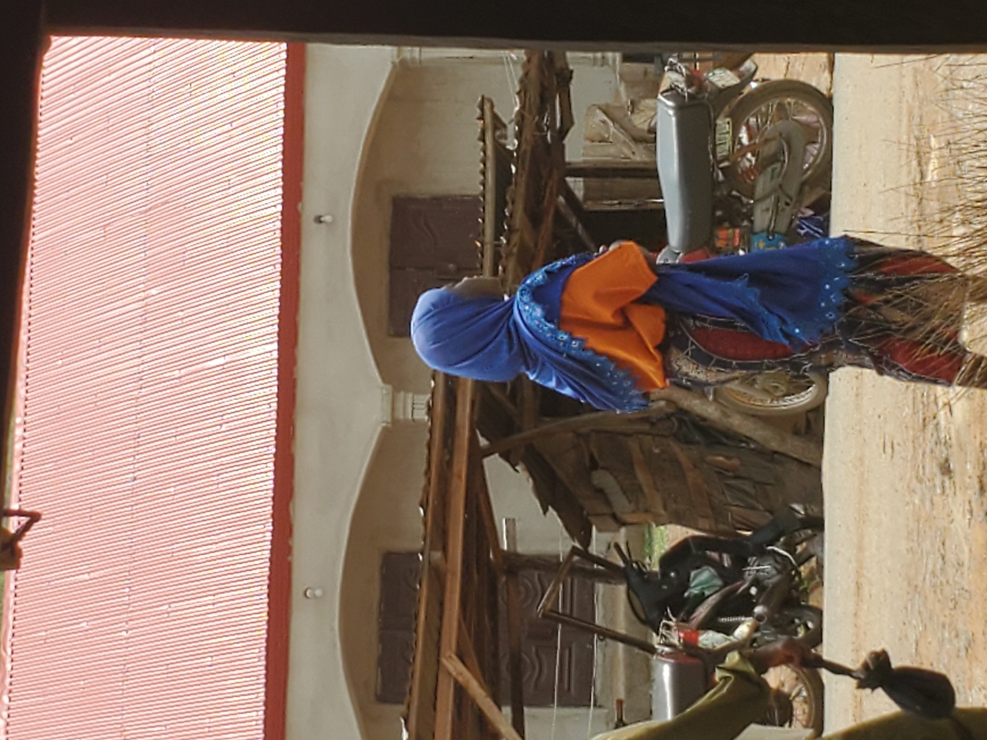 A scene in Mararaba, Nigeria, shows a woman with a blue headscarf and orange top on a motorcycle. The background reveals a building with a corrugated metal roof and arched facade. Another motorcycle is parked nearby. Wooden structures are also visible in the background. The ground appears to be dirt or concrete.