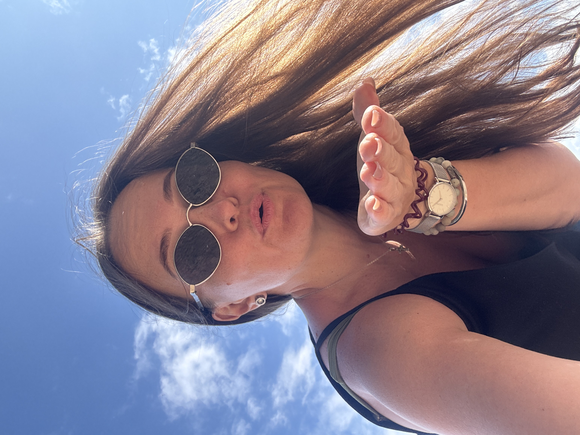 A woman with long brown hair and sunglasses is blowing a kiss towards the camera. She is wearing a black tank top and several bracelets on her left wrist, including a watch. The background is a clear blue sky with a few wispy clouds. The photo appears to be taken outdoors on a sunny day. There is no identifiable information in the image that places it in Chaikovsky, Russia.