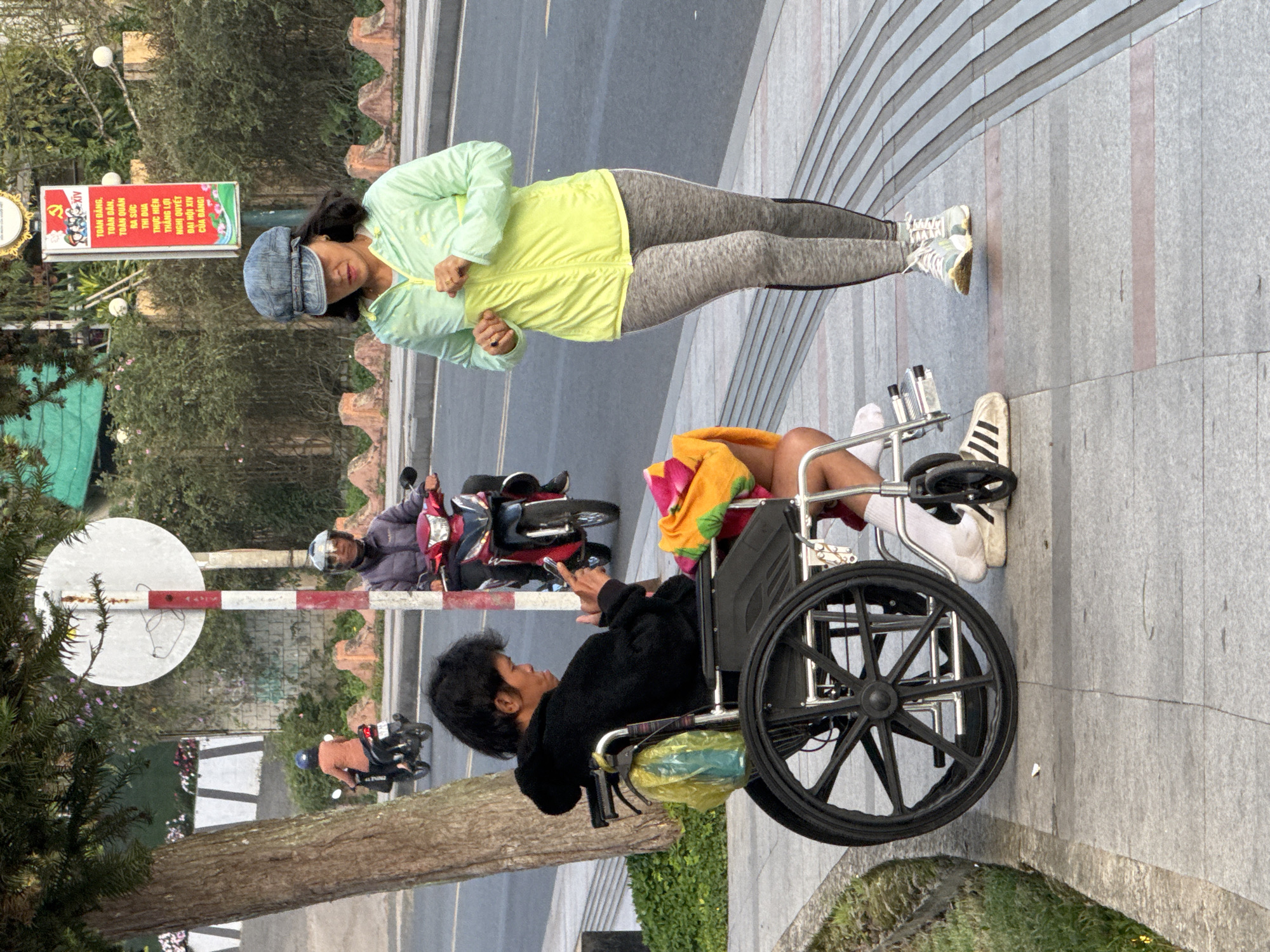 In Da Lat, Vietnam, a person sits in a wheelchair on a sidewalk next to a tree, looking at their phone. A woman in athletic wear and a hat stands on the sidewalk opposite them, facing away from the viewer and looking down at her hands. A red and white striped pole with a sign is visible behind the person in the wheelchair. In the background, two people on motorcycles are on the road. The sidewalk is made of granite pavers.