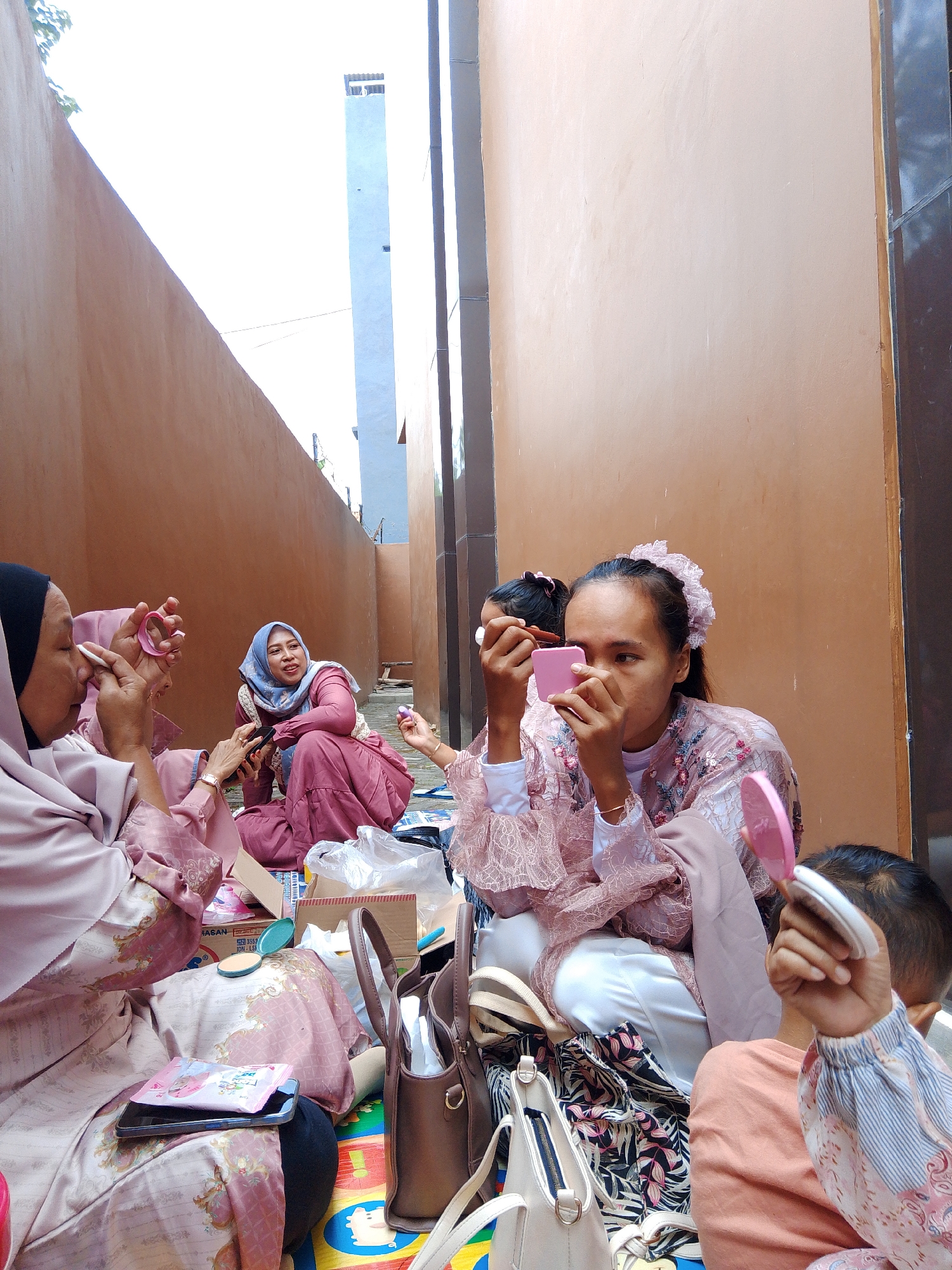 In a narrow alleyway between two ochre-colored buildings, a group of five individuals, all wearing variations of pale pink and maroon traditional clothing and headscarves, are seated on a colorful mat. They appear to be preparing for an event, as several of them are applying makeup. A woman in a black headscarf, on the far left, holds a small pink mirror and a white puff, dabbing powder on her cheek. To her right, another woman in a blue headscarf looks towards her and smiles. In the center, a young woman with a pink hair accessory is holding a pink compact mirror and applying makeup to her face. To her right, a child with dark hair is holding a small pink mirror and looking away from the camera. The ground is scattered with makeup compacts, brushes, bags, and various items of clothing. The background shows the exterior walls of the buildings, with a tall, light blue structure visible in the distance, likely part of a larger building. The lighting suggests it is daytime, with bright sunlight illuminating the scene. The location is described as being in the city of Tasikmalaya, Indonesia.