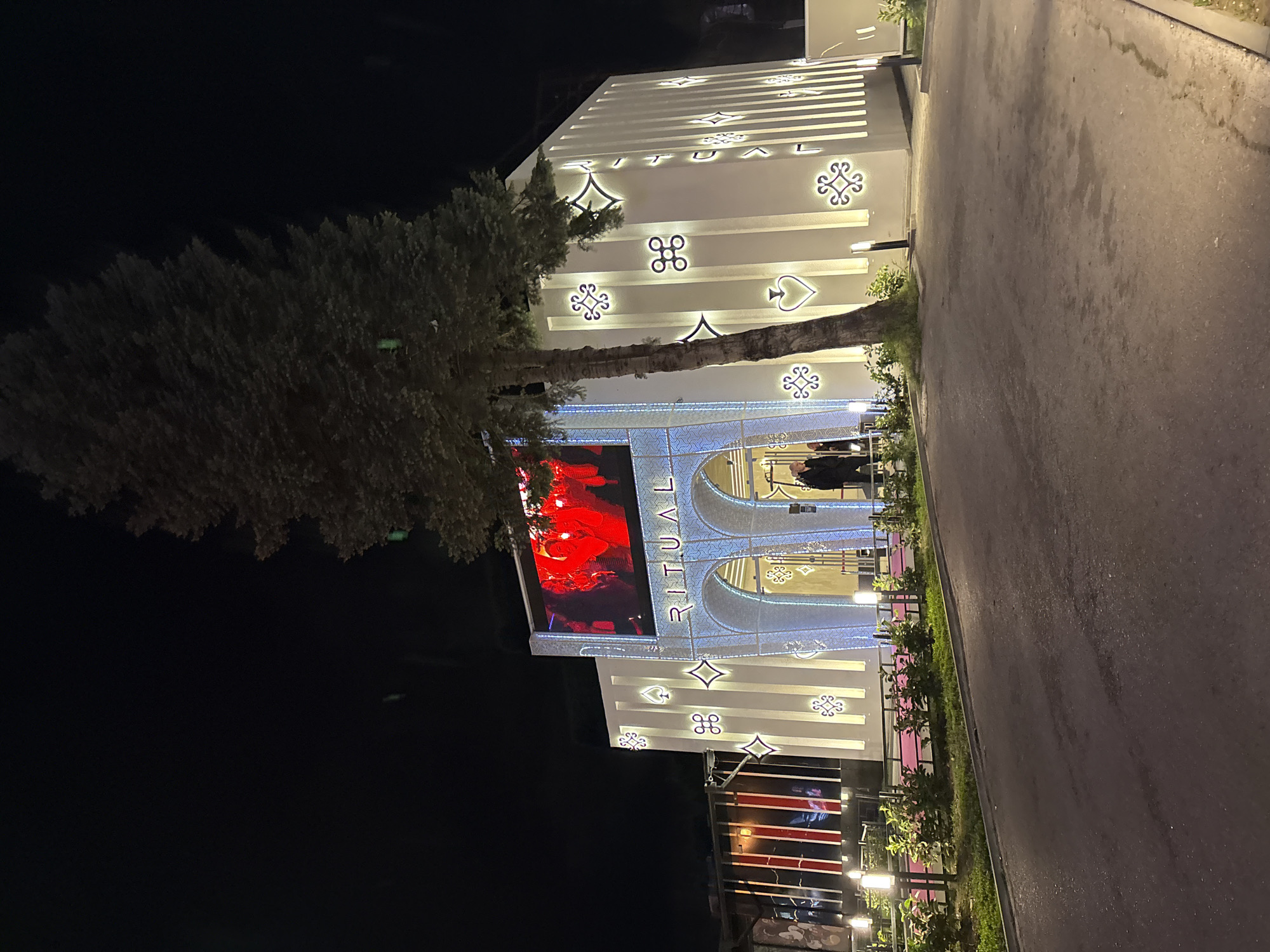 The image shows the exterior of a building at night, identified by the name "RITUAL" in vertically stacked illuminated letters above an arched entrance. The building is decorated with various symbols including diamonds, spades, and other geometric designs. A large screen displays red and white imagery. A tree is positioned near the building's facade. A paved road runs alongside the building. Sofia, Bulgaria is not mentioned in the photo.