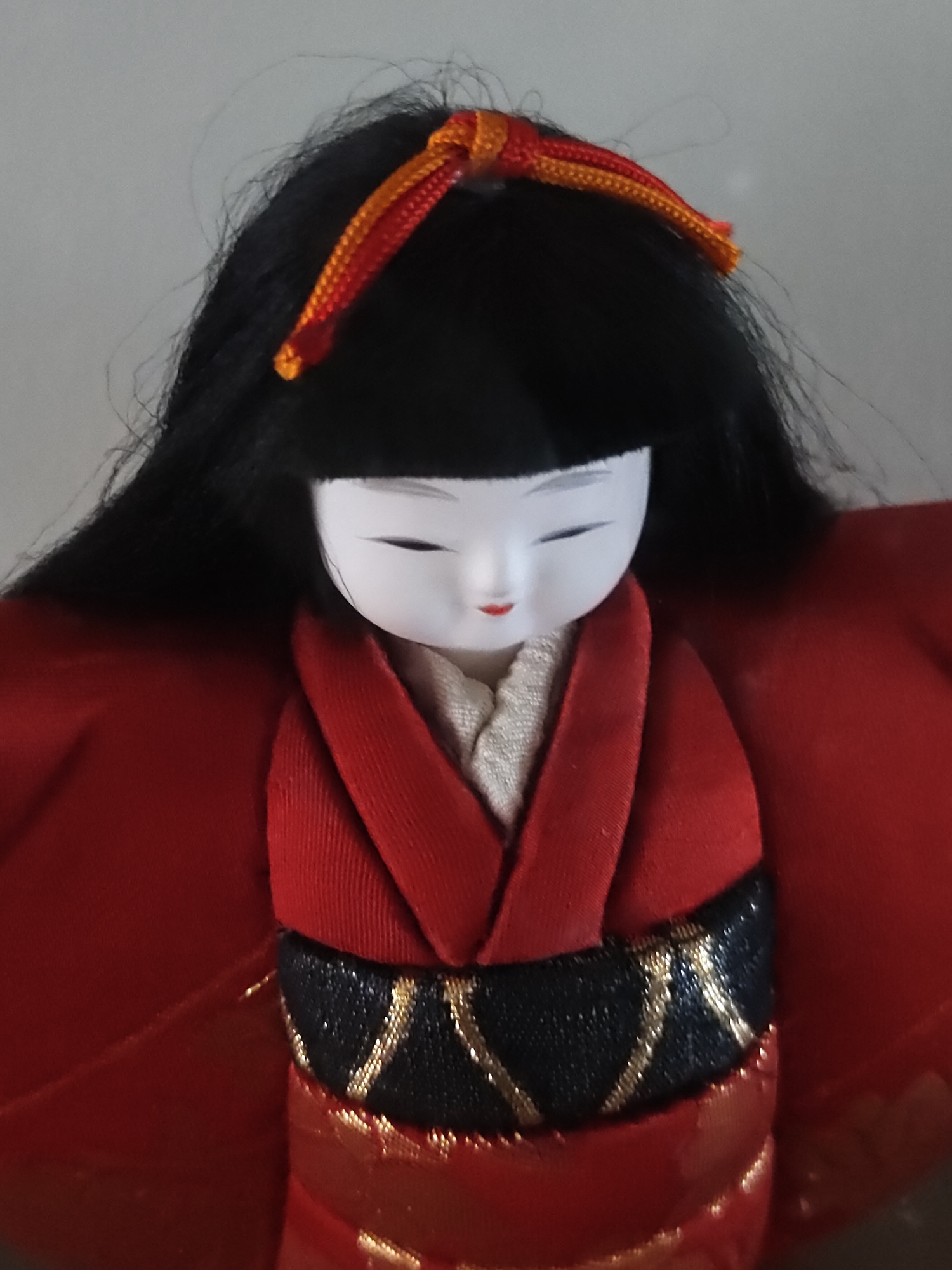 The image displays a close-up, vertical shot of a traditional Japanese doll, presented from the chest upwards against a plain, light grey background. The doll has a smooth, white porcelain-like face with narrow, downturned black eyes and small, red painted lips. Its hair is black, styled with a straight fringe across the forehead and long, flowing sides. An orange and red braided cord is tied as an ornament on the top of its head. The doll wears a red outer garment, consistent with a kimono, with visible folds and texture. Beneath the red outer layer, a lighter, cream-colored inner garment or collar is visible at the neckline. A black obi, adorned with diagonal golden patterns, cinches the waist, with further red fabric and hints of gold detailing visible below it.