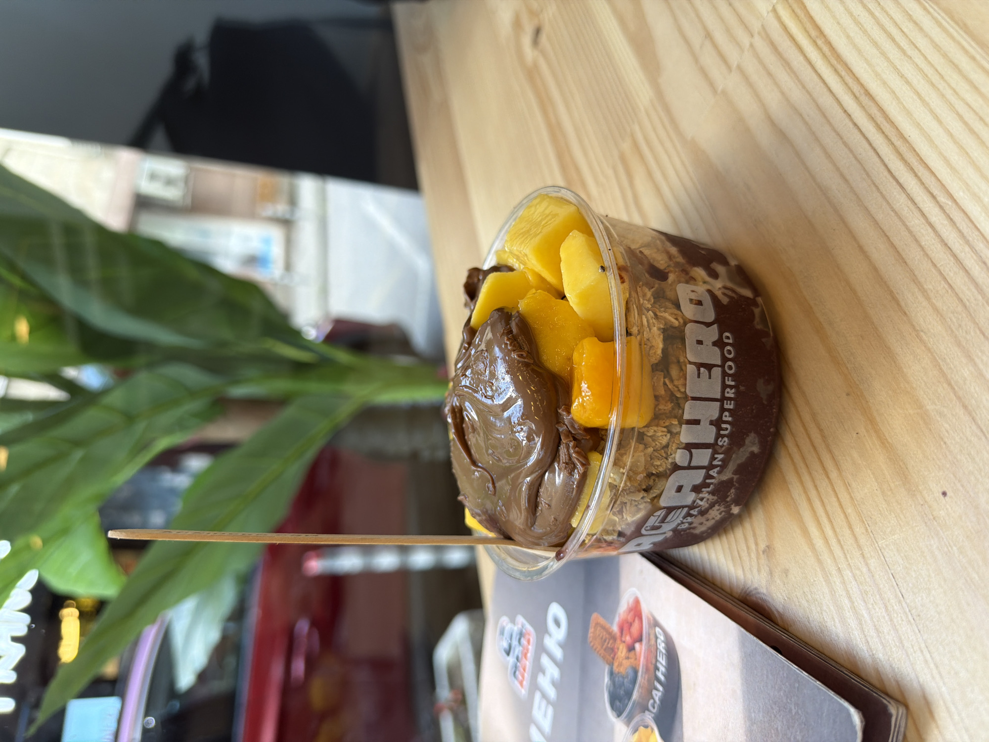 A close-up, slightly low-angle shot shows a clear plastic cup filled with acai, granola, mango chunks, and a dollop of chocolate spread, all topped with a wooden skewer. The cup is positioned on a light-colored wooden table. To the left of the cup, a folded menu with images of acai bowls is visible. In the background, out of focus, are green leaves of a plant, a red vehicle, and a building facade with windows. The lighting suggests an indoor setting, possibly a cafe or restaurant, on a bright day. The overall impression is of a refreshing and appealing food item.