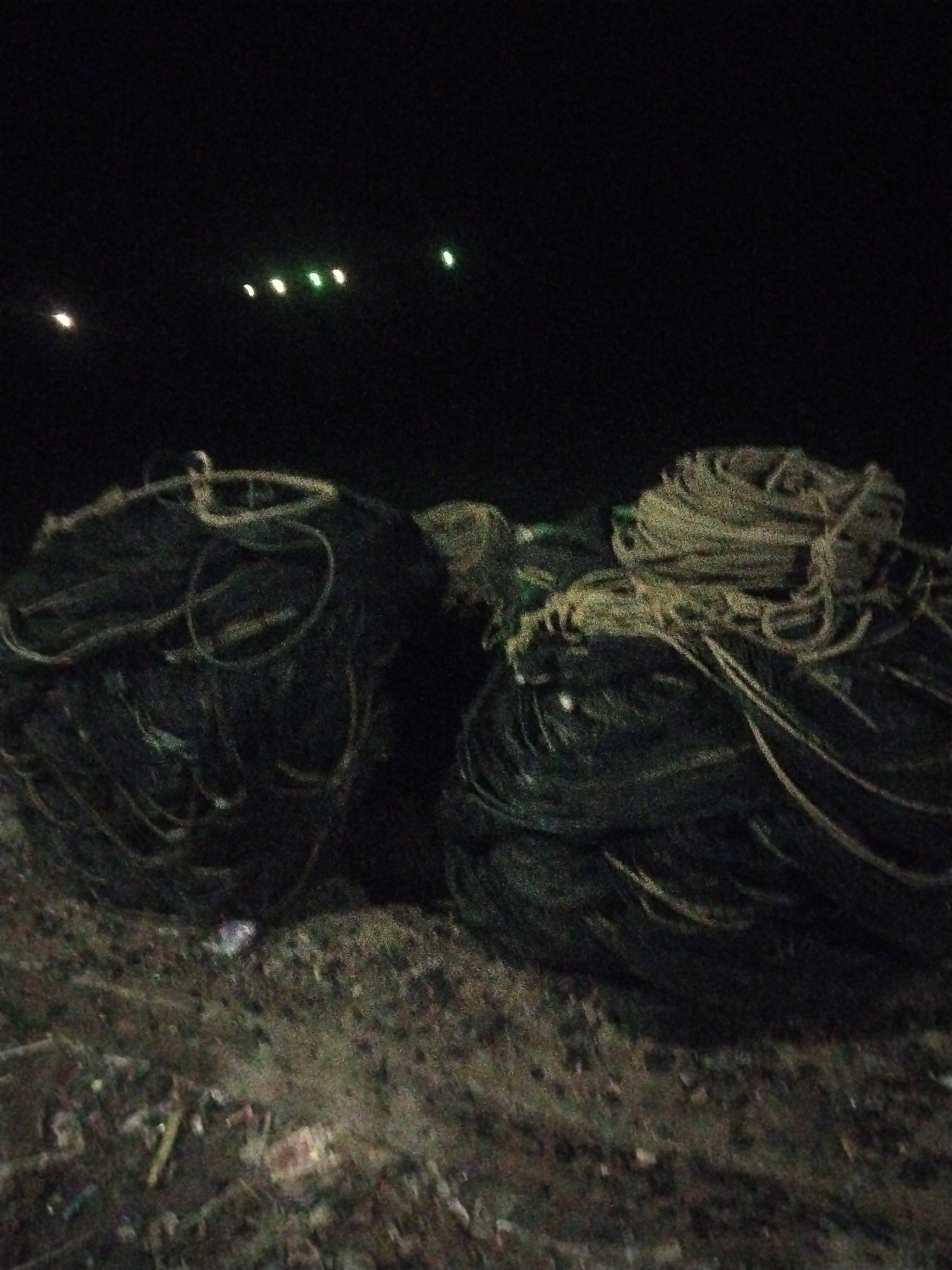 The image depicts two large, dark, circular bundles of what appear to be fishing nets or ropes. These bundles are resting on a rough, uneven surface, possibly dirt or gravel, with some debris scattered around their base. In the background, against a dark sky, there are several small, out-of-focus lights. One light is a single white or yellow point on the far left, followed by a cluster of three greenish-white lights, and then another greenish-white light further to the right. The overall scene is dimly lit, suggesting it was taken at night. The specific location is Lhokseumawe, Indonesia.