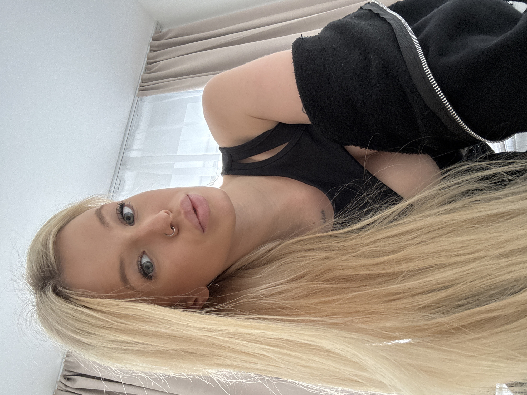 A young woman with blonde hair, blue eyes, and a nose ring is pictured from the chest up. She is wearing a black tank top and a black jacket draped over her shoulders. The background shows beige curtains and a window. There is no visible indication of Saint Petersburg, Russia in the image.