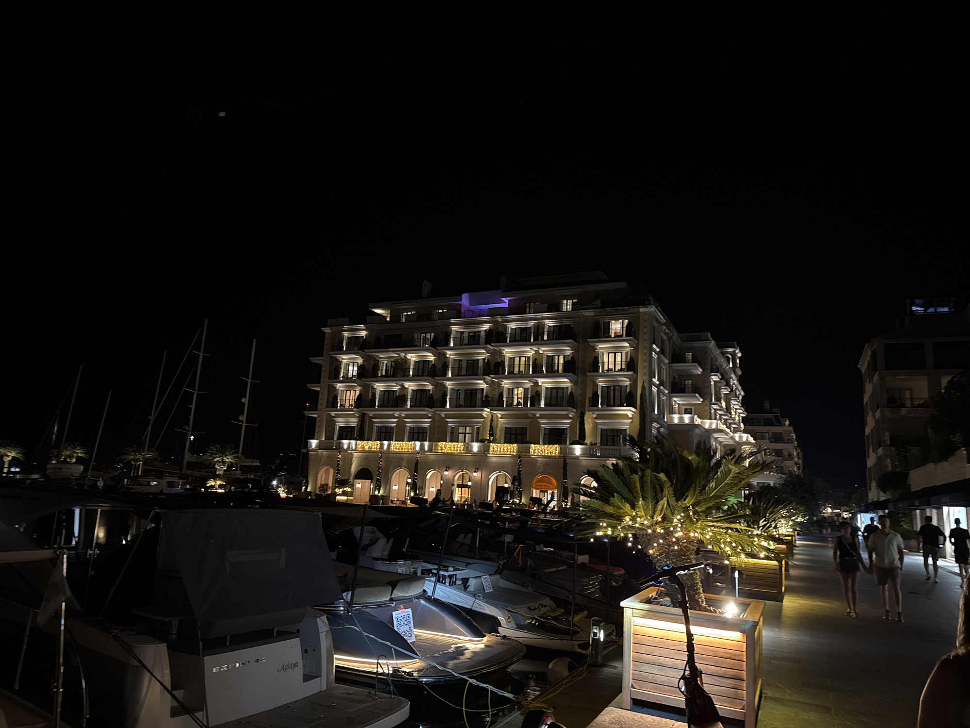 At night, a multi-story hotel building with a beige facade and numerous illuminated windows stands on the waterfront. The building is lit with warm yellow lights, and a section of the roof is illuminated with purple light. In front of the hotel, a row of moored yachts and sailboats are visible. A paved walkway runs alongside the marina, lined with planters containing palm trees decorated with string lights. Several individuals are walking along the path, some in pairs and some alone. An electric scooter is parked near one of the planters. The sky is dark and clear. The scene is set in Porto Montenegro, Tivat, Montenegro, a luxury marina and residential complex.