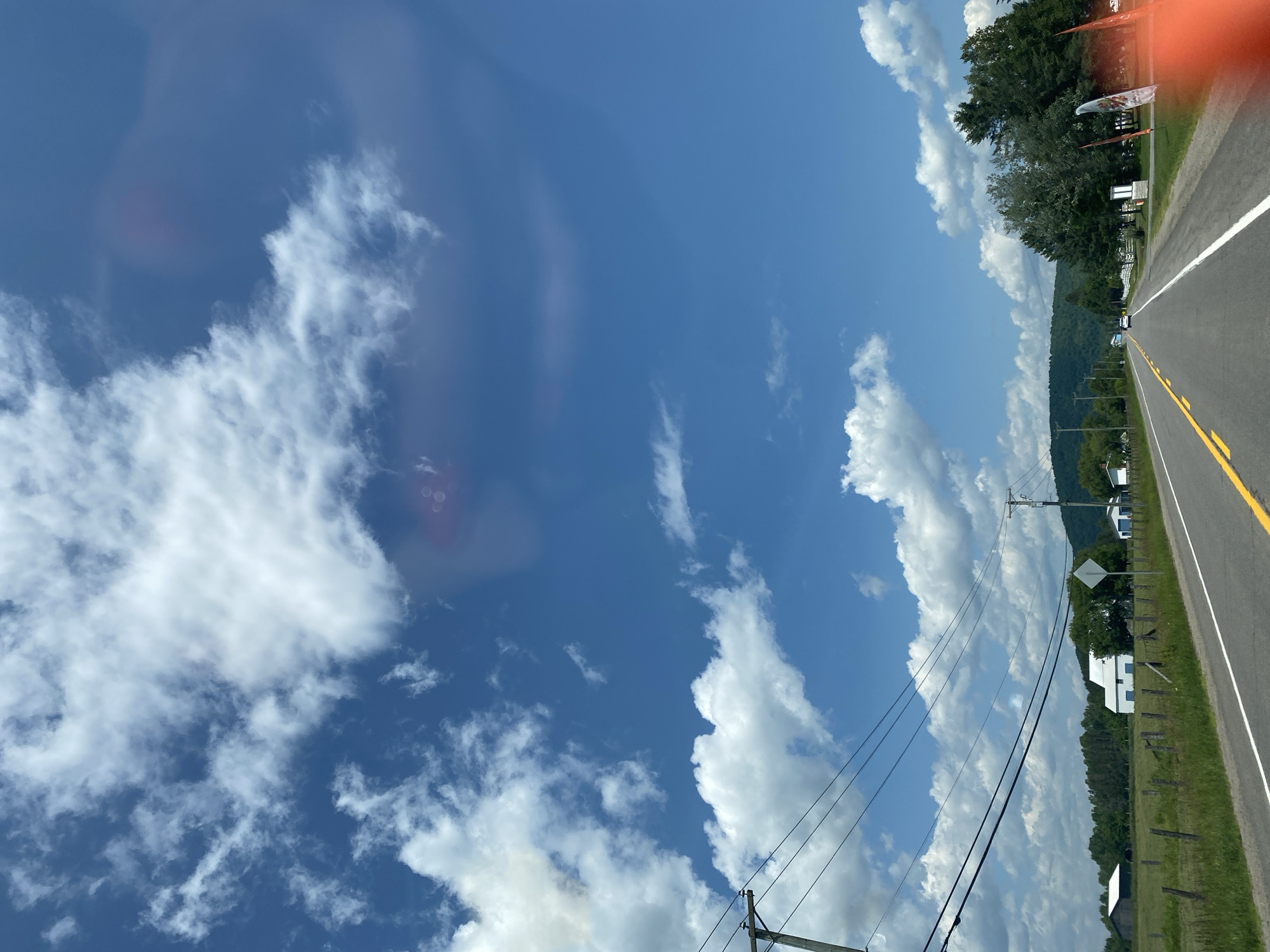 This is a vertical, medium shot of a rural road scene in Saint-André-Avellin, Canada, under a bright blue sky dotted with fluffy white clouds. The road, paved and divided by yellow lines, stretches into the distance, flanked by a grassy shoulder and a white line. To the left of the road, a wooden fence encloses a green field containing a dark barn and a white building with blue accents. Power lines run diagonally across the frame from left to right, supported by utility poles. In the background, a line of trees and rolling green hills form the horizon. On the right side of the road, several trees stand, and further down, a white fence and some orange flags are visible. The overall impression is one of a sunny, clear day in a tranquil countryside setting.