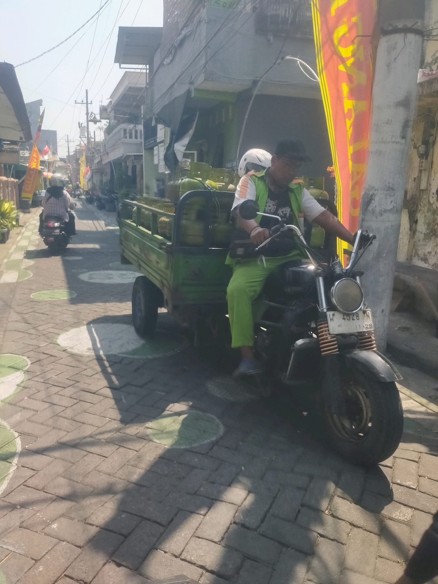 In Sidoarjo, Indonesia, a man on a black motorcycle is driving down a narrow street paved with interlocking bricks. He is wearing a green vest over a t-shirt and bright green pants, with a black hat. His motorcycle has a license plate that reads "W 4928 N 11-28". He is pulling a green, three-wheeled cart filled with what appear to be propane tanks. The cart is attached to the back of his motorcycle. In the background, another person is riding a scooter, and buildings line the street. The scene is illuminated by sunlight, casting shadows on the pavement. A large, orange and yellow banner hangs from a pole on the right side of the street.