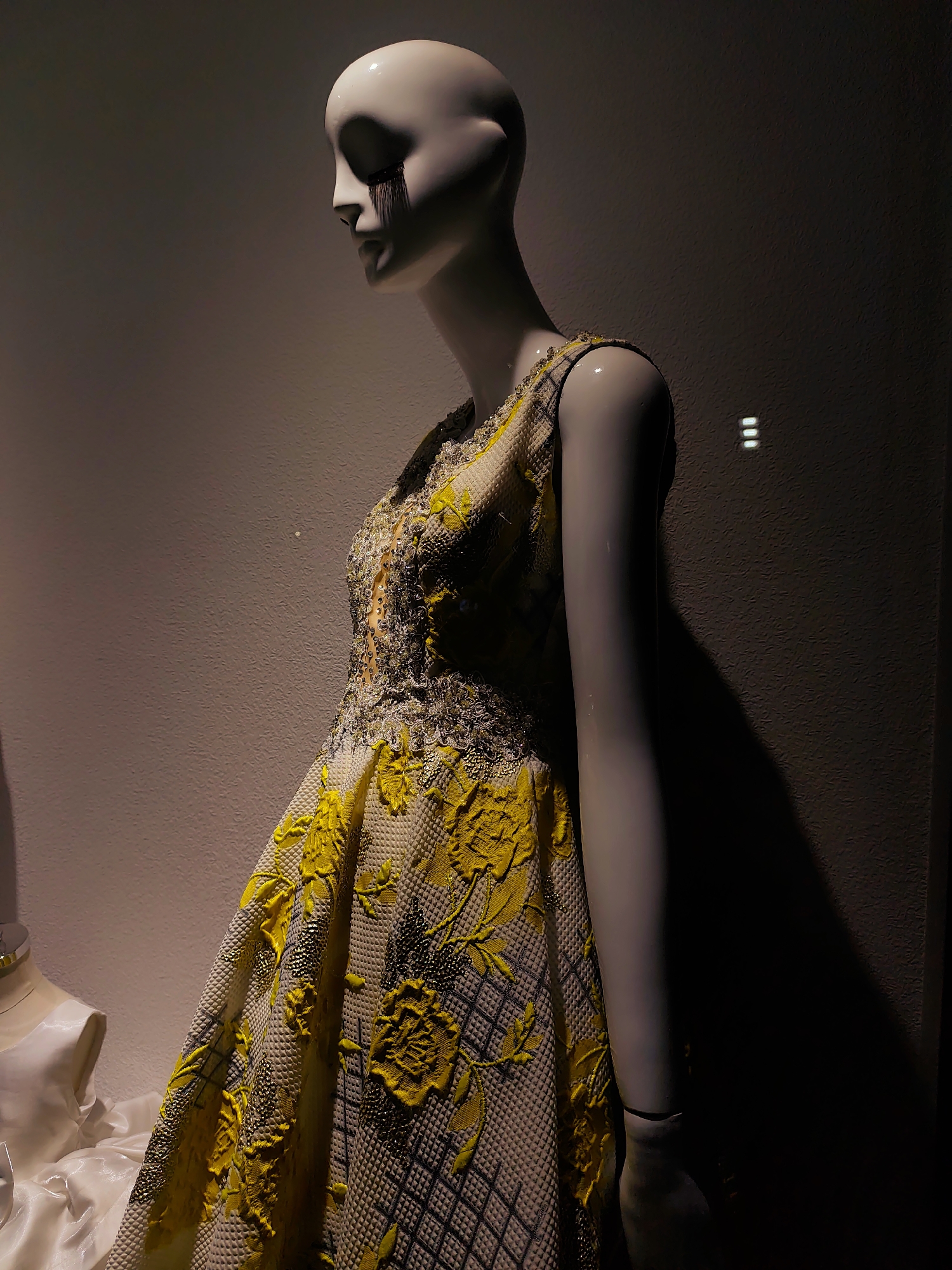 The image shows a mannequin in a window display, wearing a detailed yellow and white textured dress with floral embellishments and beading. The mannequin's head is tilted downwards, and it has black markings resembling tears or paint streaks under its closed eyes. The background is a textured wall with subtle lighting and shadows, including three bright, horizontal rectangular lights on the right side. The image is dark with the mannequin and dress illuminated. The scene is indoors, likely in a retail store. The location is not specified in the image or its context.