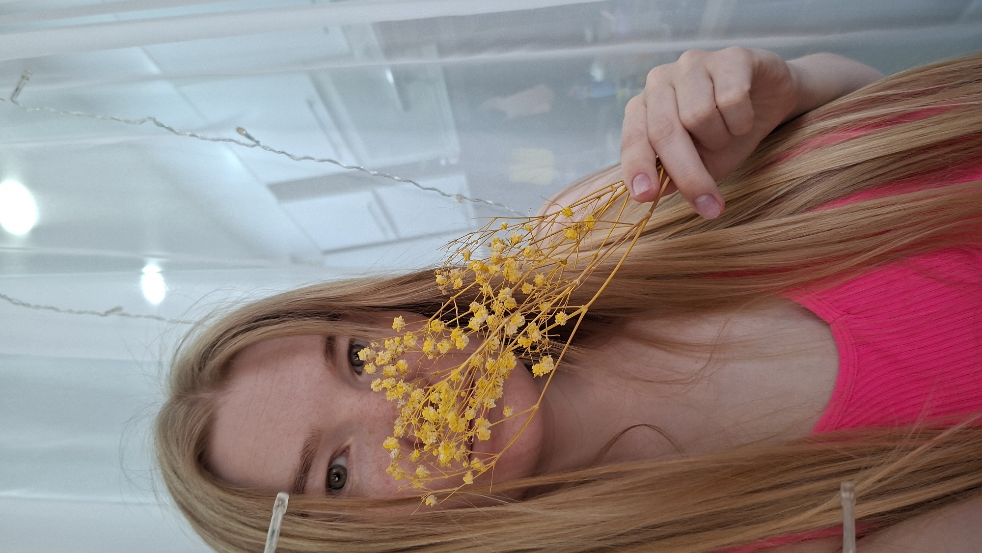 The image depicts a woman lying down, possibly indoors, with a bunch of small yellow flowers held in front of her face. She has light blonde hair and a pink top. String lights are visible in the background, possibly part of the room's decor. The location is stated to be in the city of Chaikovsky, Russia, but this cannot be confirmed by visual elements alone.
