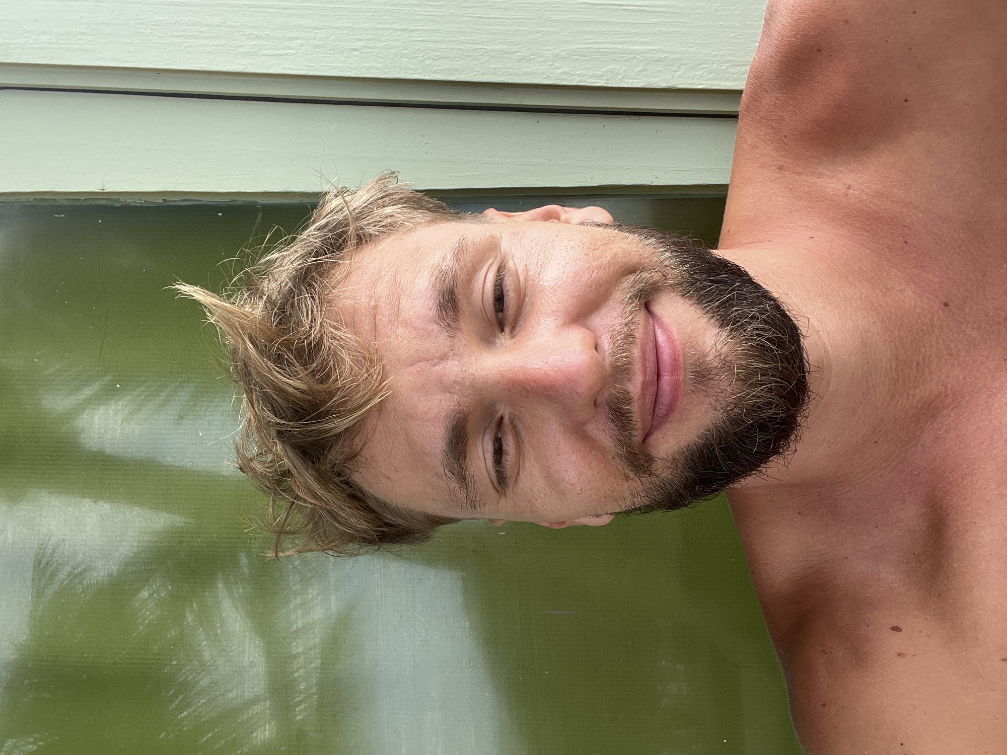 A man with blonde, messy hair and a beard smiles at the camera. He is shirtless and appears to be outdoors, with a green wall behind him that has shadows of palm leaves on it. To the right of the green wall is a light green wooden structure. The image was taken in Rajgama, Sri Lanka.