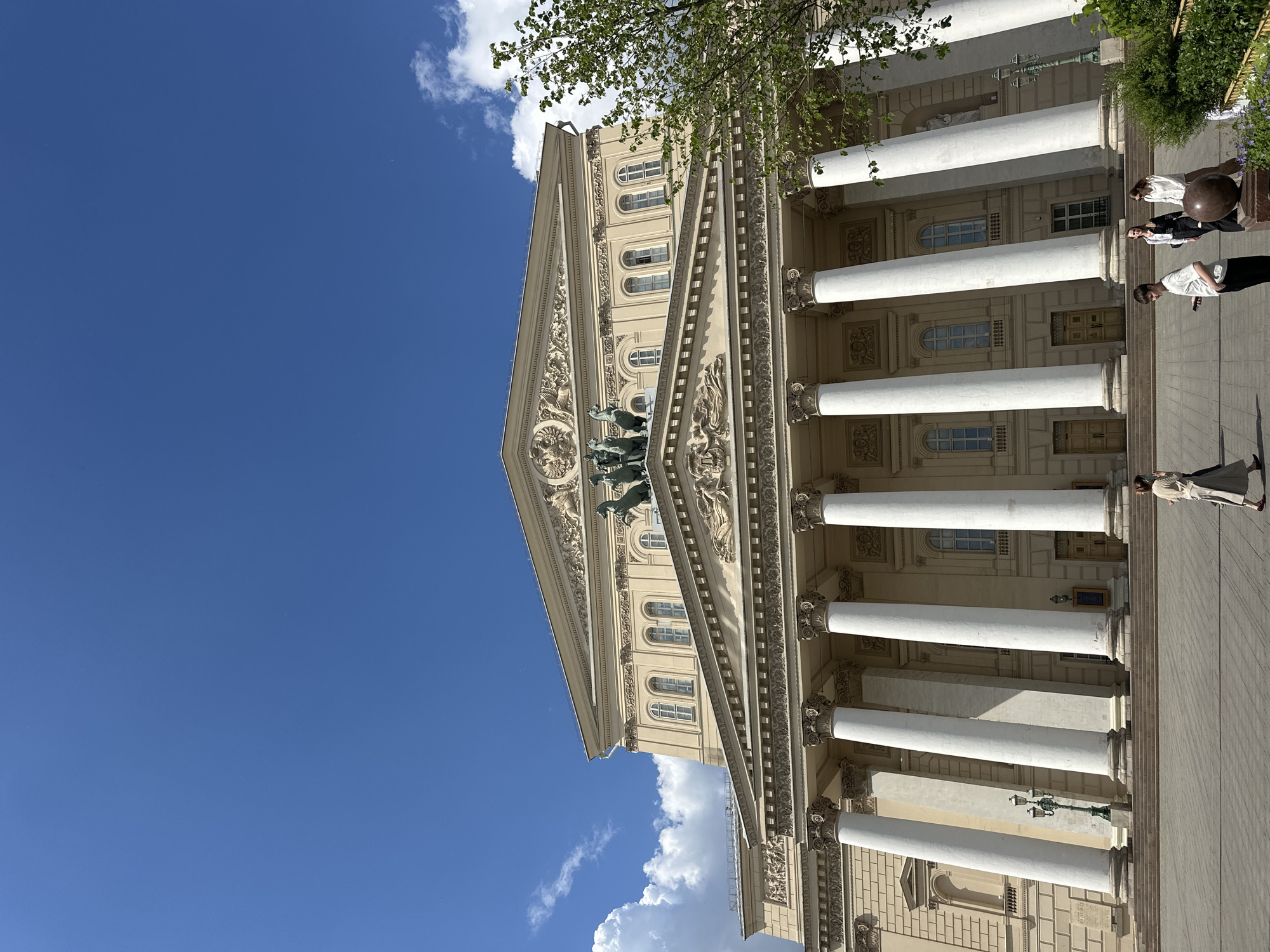 The image captures the grand facade of the Bolshoi Theatre in Moscow, Russia, presented in bright daylight under a clear blue sky dotted with scattered white clouds. The majestic neoclassical building is a light beige or yellow hue, featuring a prominent portico with six imposing white Corinthian columns. These columns support a large triangular pediment crowned by a striking bronze quadriga sculpture, depicting Apollo in his chariot pulled by four horses. Intricate classical architectural details, including ornate friezes and arched windows, adorn the building's upper levels. Green foliage from nearby trees partially frames the top right of the structure.
A wide paved plaza stretches before the theatre, where several people are seen. On the right, close to the building, two individuals are standing near a dark spherical decorative object on a low pedestal. Another person in a white shirt and dark trousers stands further back on the right. Towards the far right, a woman dressed in a light-colored coat or dress is walking away from the building. The distinct shadows cast by the columns and people indicate bright sunshine, suggesting the time of day is likely mid-day. No discernible text is visible on the building or in the immediate surroundings. The scene depicts a serene public space, with visitors casually observing or passing by a renowned cultural landmark.