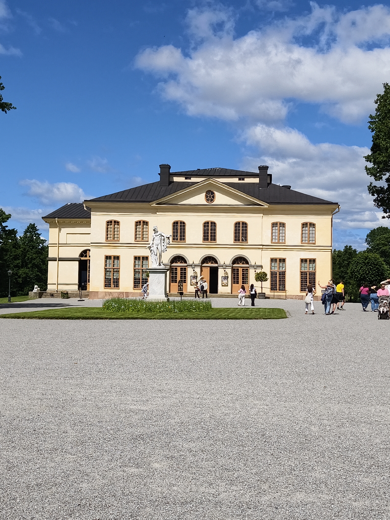 The image depicts the Drottningholm Palace, located in Ekerö kommun, Sweden. The palace is a grand yellow building with a dark roof, featuring numerous windows with wooden frames. In front of the palace, a white marble statue stands on a pedestal, surrounded by a manicured lawn and flower beds. Several people are visible on the gravel path leading to the palace, some appearing to be tourists. The sky is blue with scattered white clouds, and trees frame the scene on either side, suggesting a pleasant outdoor environment. The foreground is dominated by a wide expanse of grey gravel.