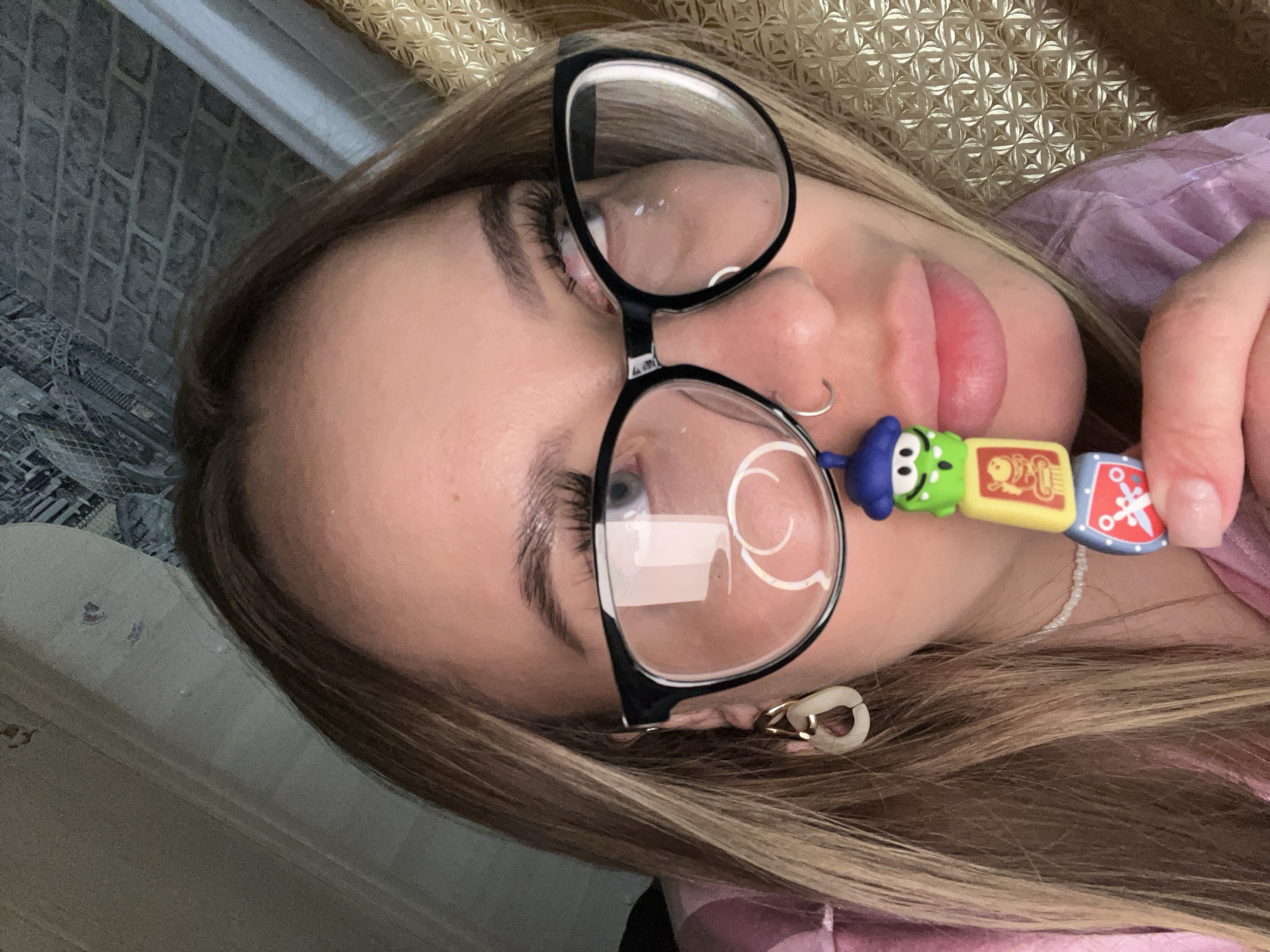 A blonde woman wearing glasses and a pink shirt is holding a colorful toy. The toy is comprised of several blocks, each decorated with different designs. The woman's hair is blonde and styled with highlights. She also has a nose piercing and is wearing earrings. The background appears to be a wall with a textured pattern. The location of the image is in the city of Novopokrovskaya, Russia.