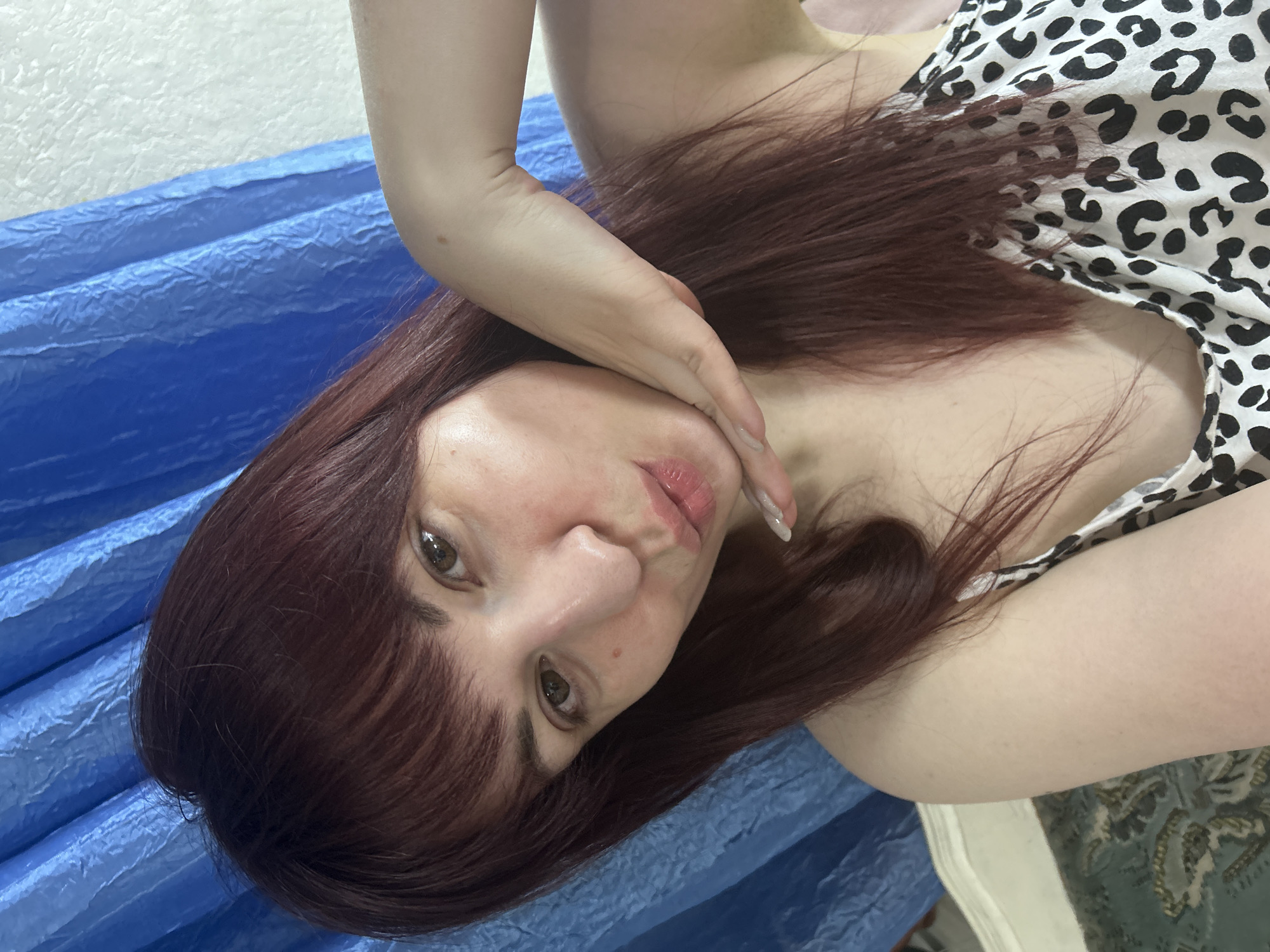 A woman with reddish-brown hair and brown eyes is posing for a close-up photograph. She has fair skin and is wearing a white top with a black animal print pattern. Her left hand is raised near her face, with the fingertips gently touching her cheek. The background consists of a textured blue surface on the left and a patterned surface on the right. There is no indication of the location being Фоки, Russia, or any other specific city.