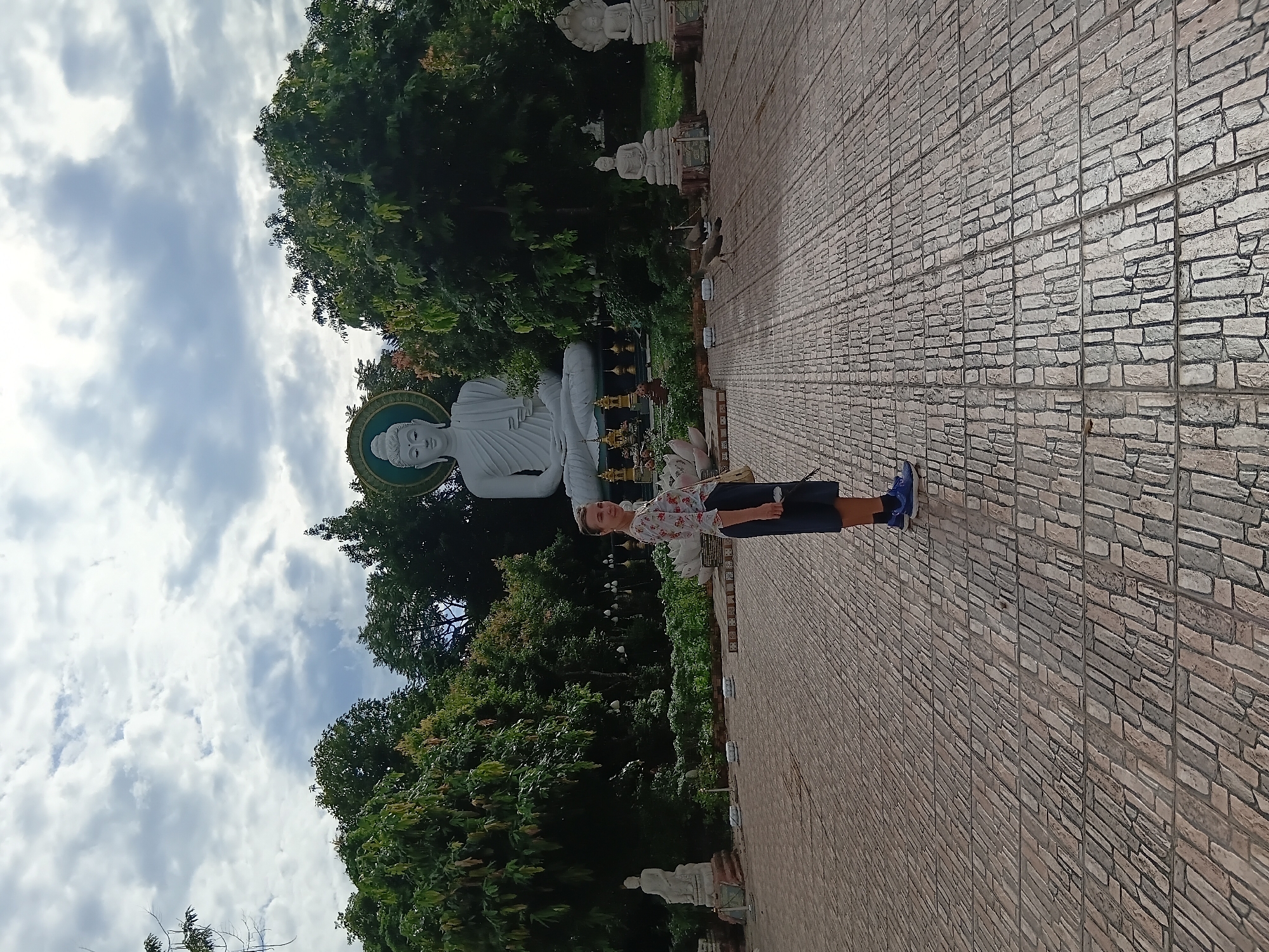 In Ban Ruammit, Thailand, a young woman with short, blonde hair stands on a paved courtyard. She is wearing a short-sleeved floral print shirt and dark shorts, with blue athletic shoes. A light-colored purse hangs over her left shoulder. Behind her, a large white Buddha statue sits in front of lush green trees and a cloudy sky. To the left of the Buddha statue, a smaller white seated Buddha is visible. To the right, there are more small Buddha statues and more trees. The courtyard is paved with rectangular stones arranged in rows.