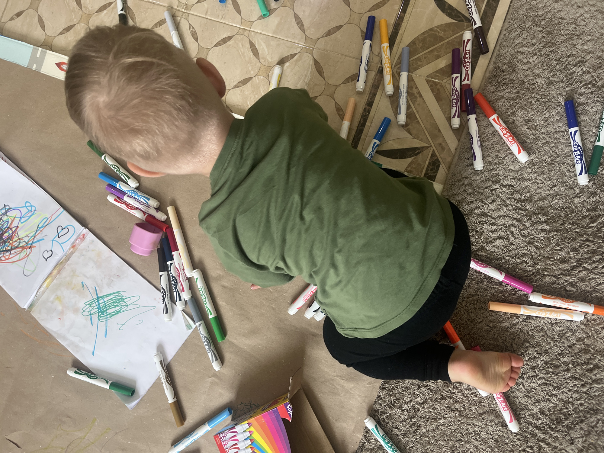 A young Caucasian child, seen from behind, is sitting on the floor surrounded by numerous colorful markers and a children's drawing pad. The child is wearing a green long-sleeved shirt and black leggings. The drawing pad is open on the floor, with scribbles in various colors, including blue and green. Several markers are scattered around the child, with some lying on a brown paper-covered floor and others on a carpeted area. The markers are of different colors and brands, predominantly "Cra-Z-Art". In the background, a tiled floor is visible. The overall scene suggests a child engaged in drawing or coloring activities. No specific location within the city of Onokivtsi, Ukraine, is discernible from the image.