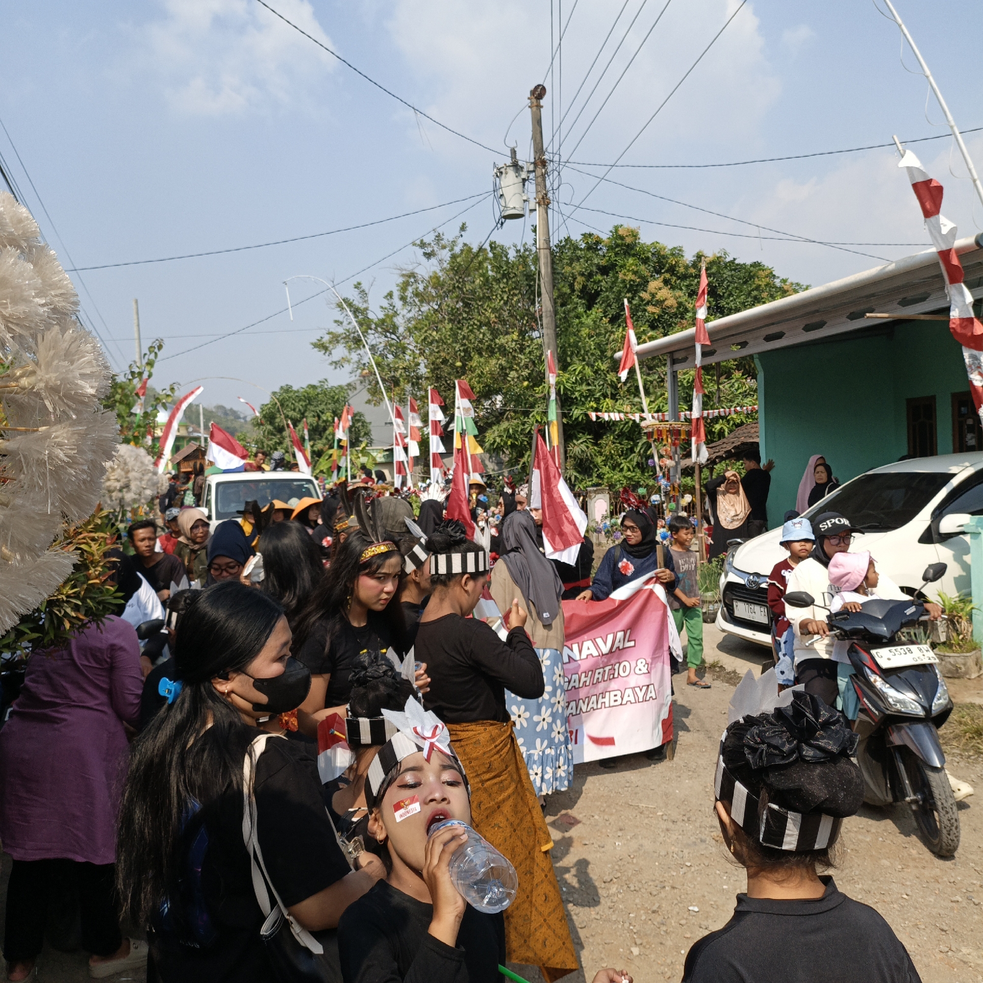 The image depicts a street scene in Tanahbaya, Indonesia, likely during a festive event or parade. A group of people, many dressed in black shirts and some wearing traditional headwear with black and white stripes, are gathered. Several individuals are holding Indonesian flags, which are red and white, and some are also carrying a banner with text that appears to read "NAVAL" and "TANAHBAYA". The atmosphere suggests a celebration, with many participants, decorations, and a general sense of community gathering. In the background, there are houses, trees, and utility poles, typical of a residential area. A white car and a motorcycle are also visible, indicating the presence of vehicles on the street. The sky is clear and blue. The focus is on the people and the celebratory elements.