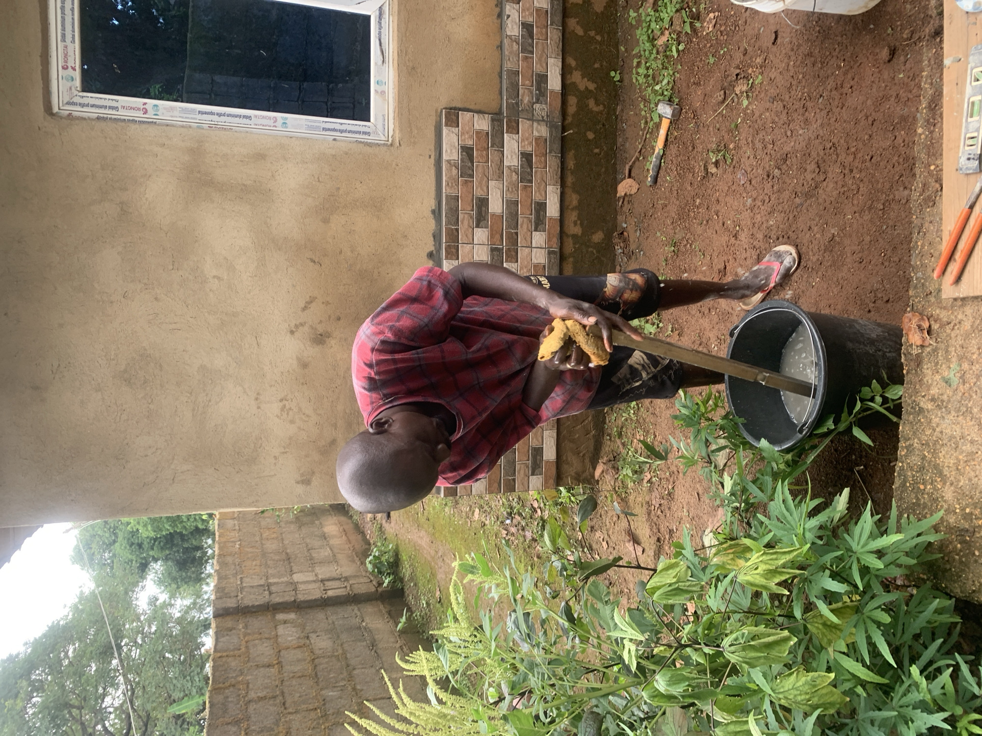 A man is standing in front of a building under construction in Agban, Nigeria. He is wearing a red and black plaid shirt and dark shorts. He is holding a wooden stick with a sponge attached to it and appears to be mixing something in a black bucket. The building has a tan-colored exterior and a window with a white frame. There is a wall made of concrete blocks to the left of the building, and vegetation is growing around the scene. Tools, including a hammer and level, are visible on the ground. The ground is covered in red dirt.