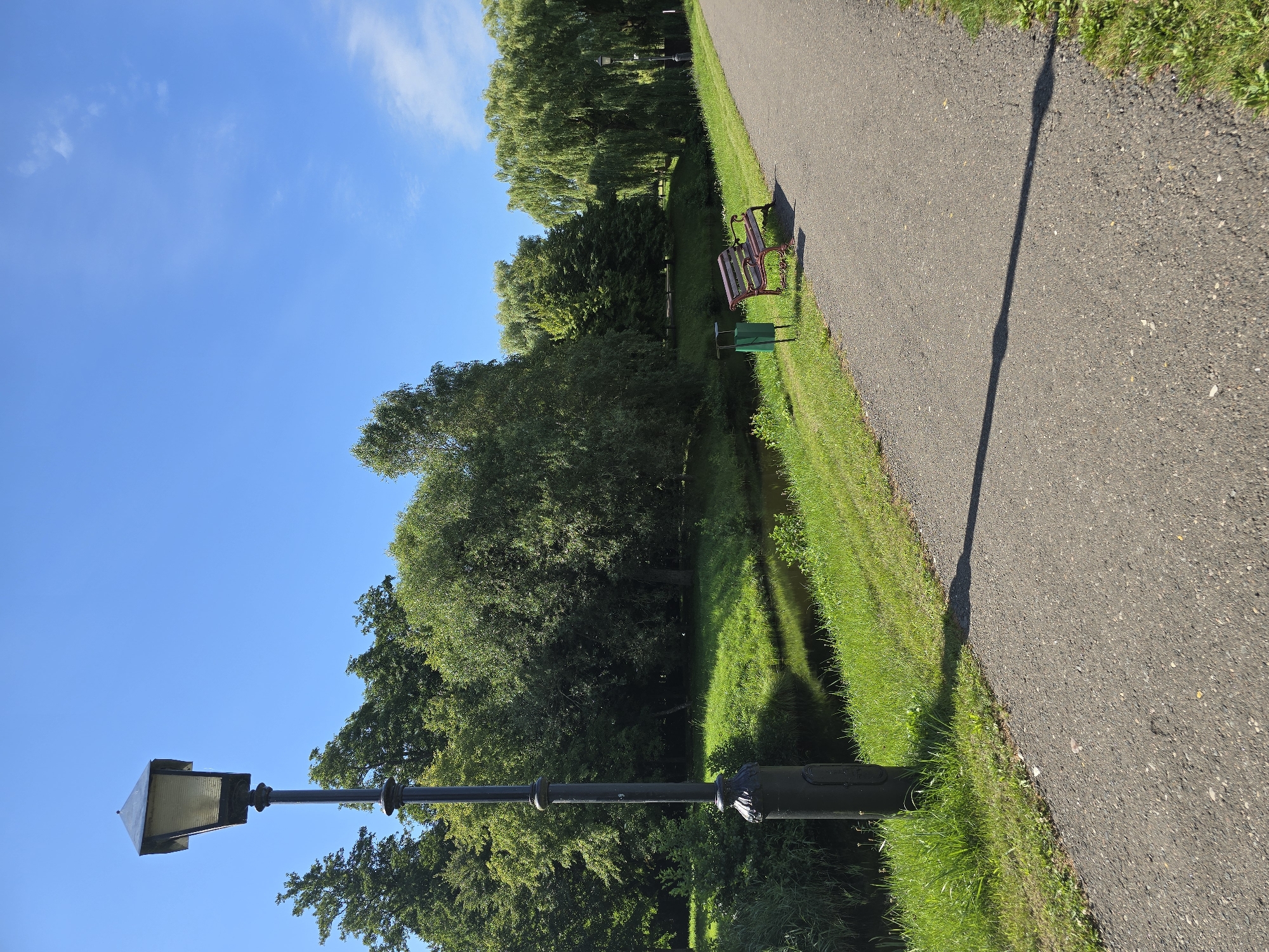 An asphalt path runs diagonally across the frame from the lower right to the upper left. To the left of the path is a strip of green grass, and beyond that is a narrow body of dark water reflecting the lush green trees on its banks. A black lamppost with a square lantern stands in the left foreground, casting a long shadow across the path. On the grass, a red and brown park bench sits next to a green trash can, and beyond that, a dense line of deciduous trees forms the background. A bright blue sky with a few wispy clouds completes the scene. This image was captured in the Niasvizh Castle park, Niasvizh, Belarus.