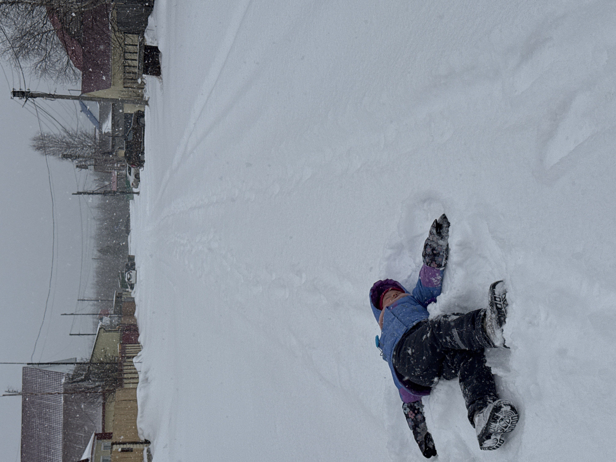 This media file captures a serene winter scene, likely in Vysoky, Russia, featuring a child playing in deep snow. The image is oriented with the street and background buildings appearing vertically along the left side, and the expanse of snow, with the child, occupying the majority of the frame.
The primary subject is a young child, dressed in warm winter clothing, lying on their back in a thick blanket of fresh snow. They are wearing a blue and purple hooded jacket, dark snow pants, winter gloves, and a matching purple and pink hat. Snow is visible clinging to their clothing, suggesting it is actively snowing. Their posture indicates they might be making a snow angel or simply enjoying the soft, deep snow.
The scene is dominated by snow, which covers the ground entirely, extending into the distance where faint tracks can be seen. In the background, a residential area is visible, with several houses featuring pitched roofs, also laden with snow. Bare trees stand along the side of what appears to be a street or path, and numerous utility poles with power lines stretch into the cloudy, gray sky. A few snow-covered cars are partially visible further down the road.
The weather is clearly snowy and cold, with small, white snowflakes distinctly visible falling through the air. The time of day appears to be daytime, but overcast due to the heavy snow conditions. There is no visible text in the image. The overall impression is one of peaceful winter play amidst a snow-covered landscape.