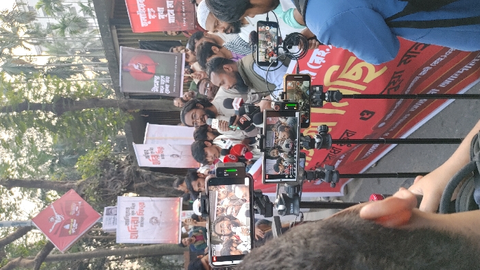 The scene depicts an outdoor press conference or public gathering in an urban area of Dhaka, Bangladesh. A central figure, a man with dark hair and a beard wearing a light-colored tunic, is speaking into multiple microphones held by various media organizations, while also holding a smartphone in his left hand. He is surrounded by a crowd of approximately 20-30 other men, some attentively listening.
In the immediate foreground, three smartphones mounted on black tripods are recording the speaker, displaying live video feeds of the event. The perspective is from behind one of these recording setups, with a portion of a person's head and shoulder visible in the lower left.
The background features several trees with visible root systems and sparse foliage, beyond which multi-story buildings are partially visible. Various protest banners are displayed:
* A red, diamond-shaped banner hangs from a tree in the upper left, featuring a white silhouette of a figure with outstretched arms above symbols resembling books and other objects, with Bengali text.
* Below it, a white rectangular banner with red text is visible.
* Further right, a dark banner shows a stylized red figure against a black background, with Bengali text and the English phrase "why shed blood on the border."
* A large red banner with prominent Bengali text stretches horizontally across the lower part of the scene, partially obscured by the tripods.
The event appears to be a politically charged gathering, given the nature of the banners and the formal press conference setup. The setting is a public street or plaza in Dhaka.