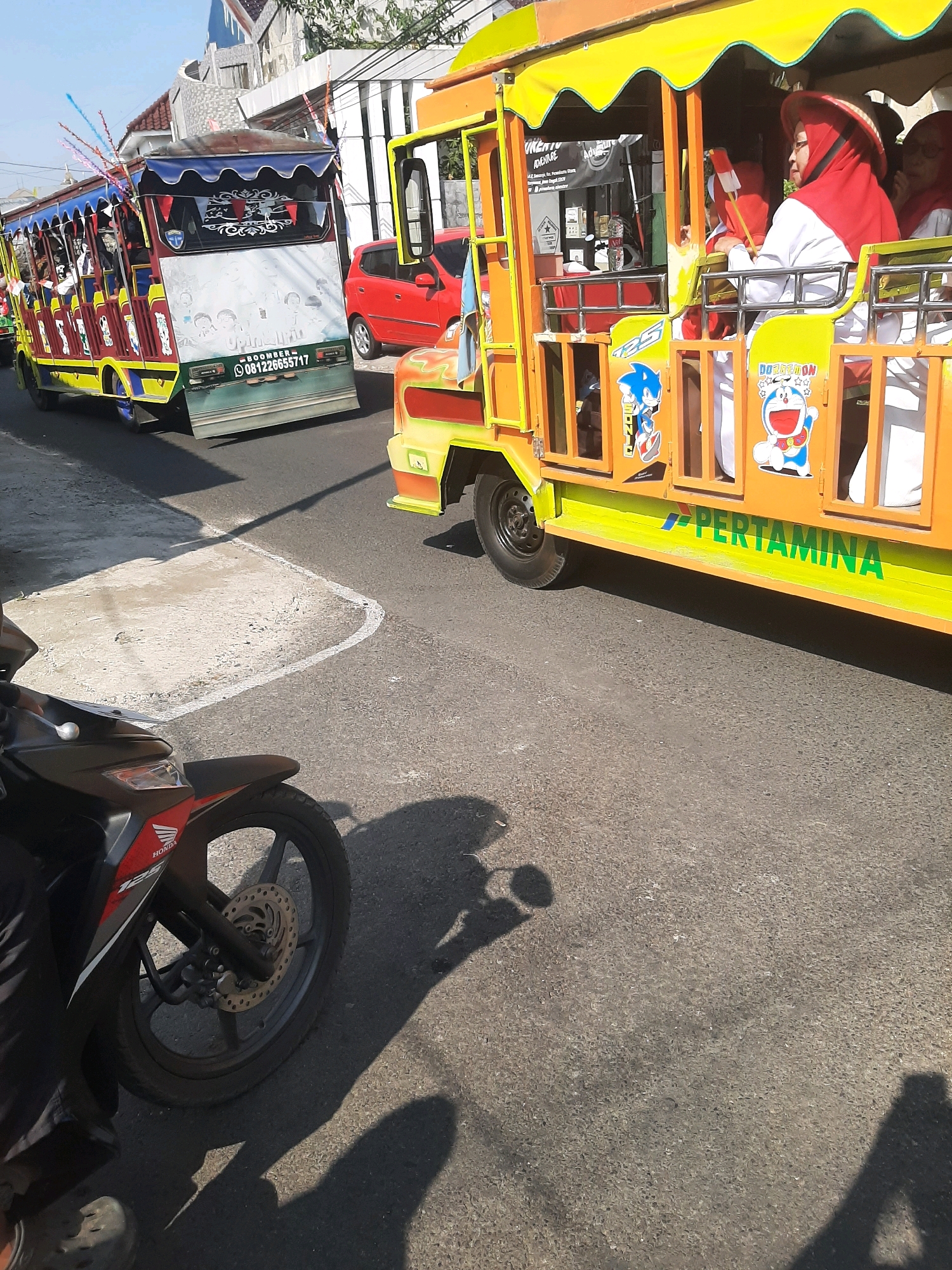 In Sumampir, Indonesia, a vibrant, open-sided yellow and orange bus, adorned with cartoon characters like Doraemon and Sonic, transports passengers along a paved road. The bus has "PERTAMINA" written in green along its side. To its left, a similar, smaller bus, primarily red and yellow with a blue canopy, is also visible, featuring a digital display showing "BOOMBER- 081226655717". A red car is parked on the side of the road. In the foreground, the front wheel and handlebars of a black and red Honda motorcycle are prominent. The scene is set against a backdrop of buildings and under a clear blue sky.