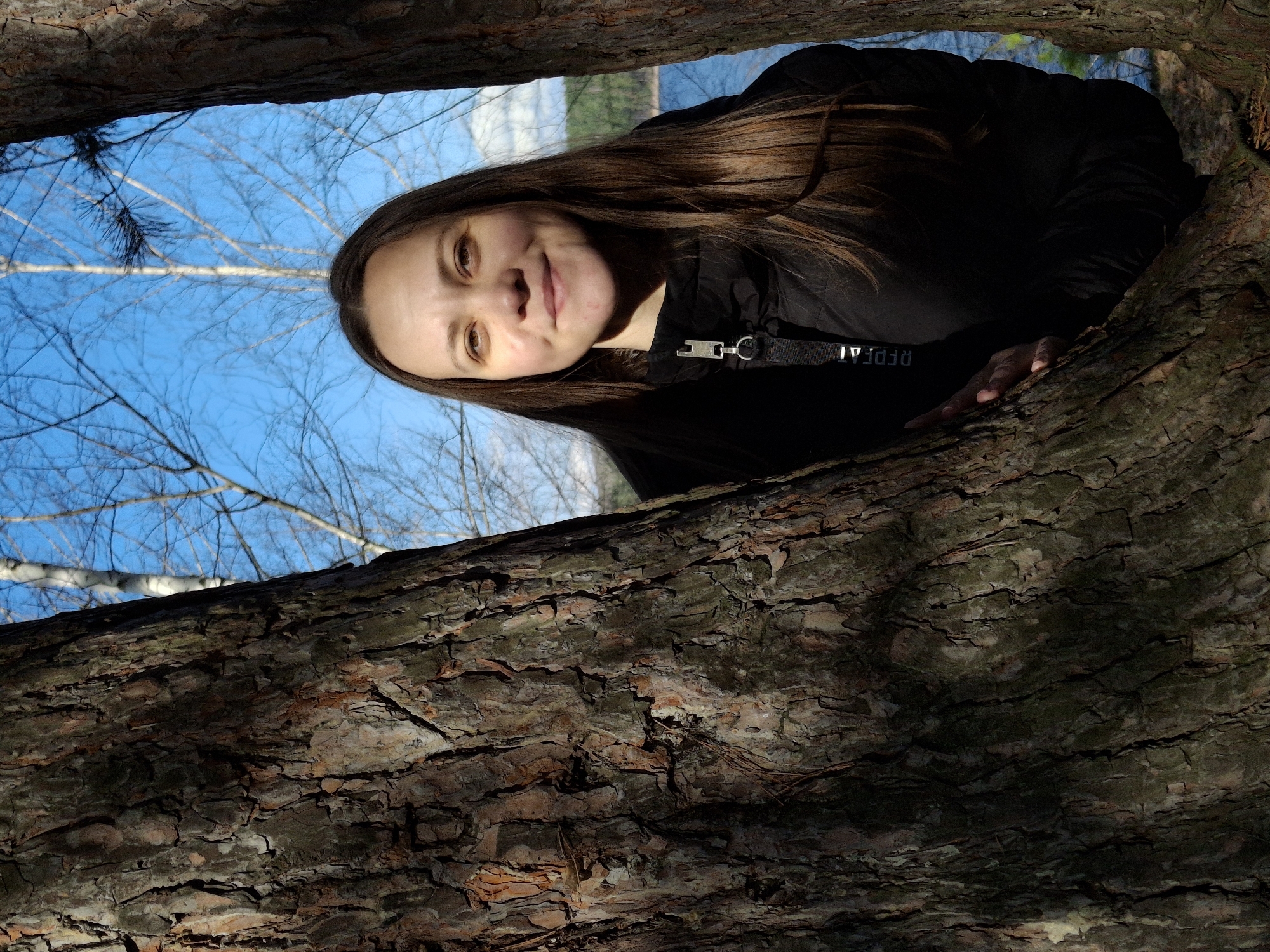 A close-up, low-angle shot captures a young woman looking directly at the camera, framed by two large, textured tree trunks. The image appears to have been taken from below, looking up through a gap in a tree or between two adjacent trees.
The woman, with long brown hair, is positioned in the upper portion of the frame. She has light skin and a gentle smile, with her eyes slightly narrowed. She is wearing a black jacket with a prominent silver clip and a black strap on which the white text "REPEAT" is visible, oriented upside down from her perspective but readable when the image is corrected. Her right hand is visible, resting on the rough bark of the tree trunk that frames her from the bottom right.
In the background, a clear blue sky is visible, interspersed with the bare branches of deciduous trees, suggesting late autumn or early spring. Hints of water, likely a lake or river, can be seen in the far distance through the opening in the trees, particularly behind her head. The tree trunks framing the scene are thick and deeply furrowed, displaying various shades of brown and green moss, with some reddish patches. The lighting suggests a bright, sunny day. The scene takes place in a natural, wooded environment, consistent with the location context of Chaikovsky, Russia.