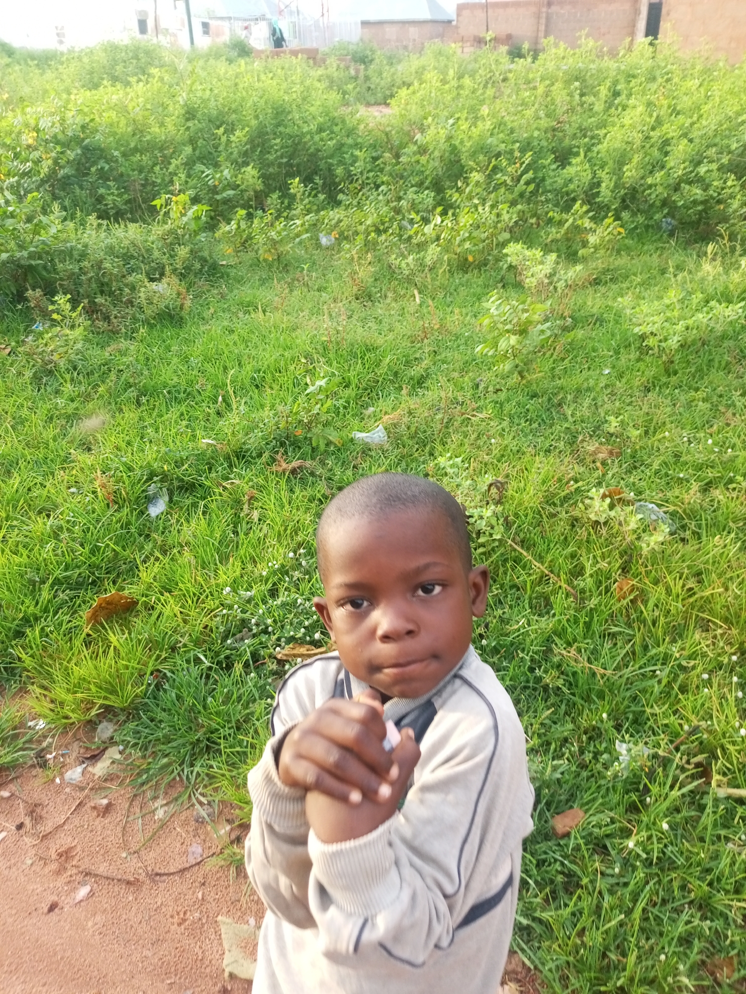In Tempure, Nigeria, a young child with dark skin and hair is positioned in the foreground, looking directly at the camera. The child wears a light-colored sweater with dark trim. Their hands are clasped together in front of their chest. The background consists of a densely vegetated area with lush green grass and various plants, suggesting an outdoor setting. Beyond the vegetation, a brick wall and part of a building are visible. The ground in the lower left of the frame is a reddish-brown dirt path.